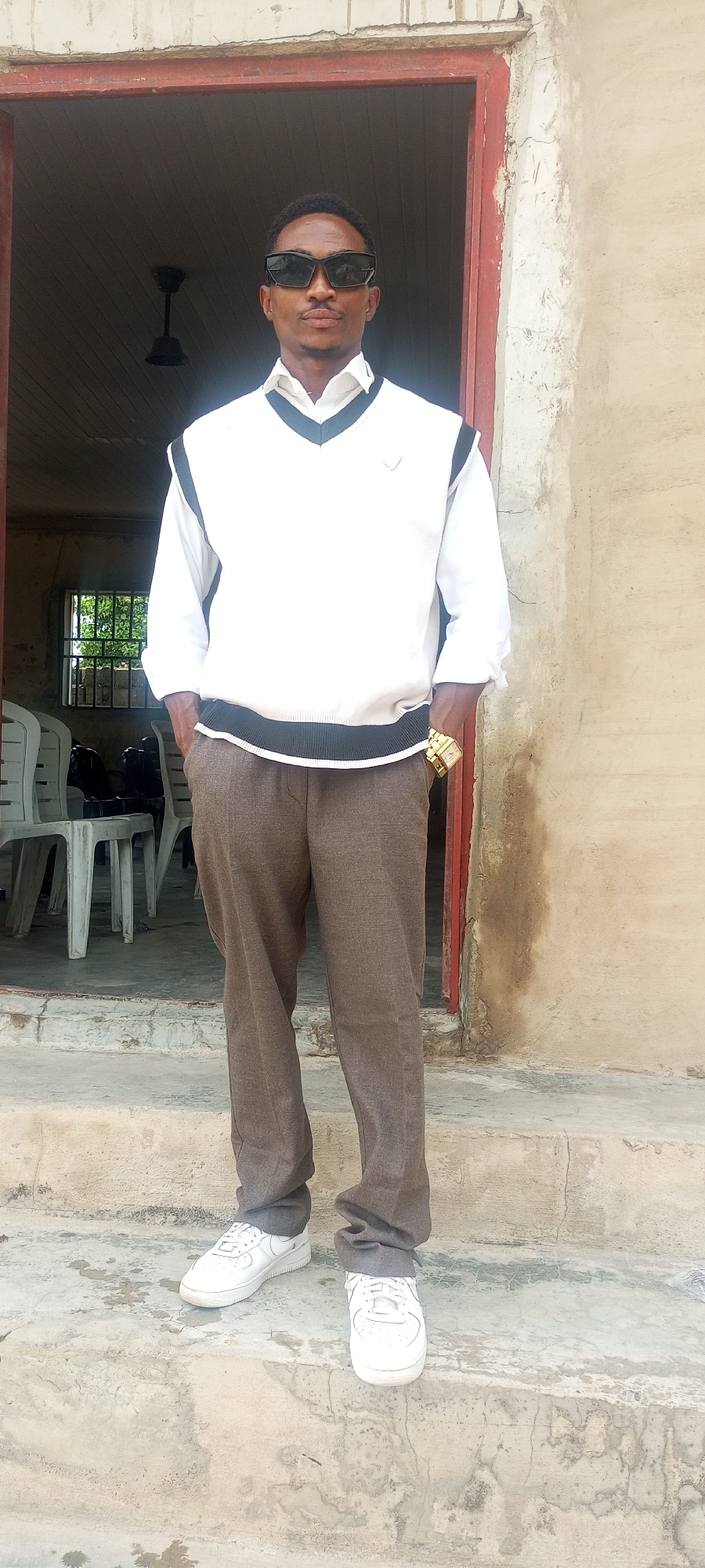 A man stands on a concrete step in front of a doorway. He wears sunglasses, a white long-sleeved shirt, a white sweater vest with black trim, brown pants, and white sneakers. He has a gold watch on his left wrist and his hands are in his pockets. Behind him, through the doorway, is a room with white plastic chairs and a barred window. The walls adjacent to the doorway are aged concrete. The location may be in Lokoja, Nigeria, but is indistinguishable and not apparent.