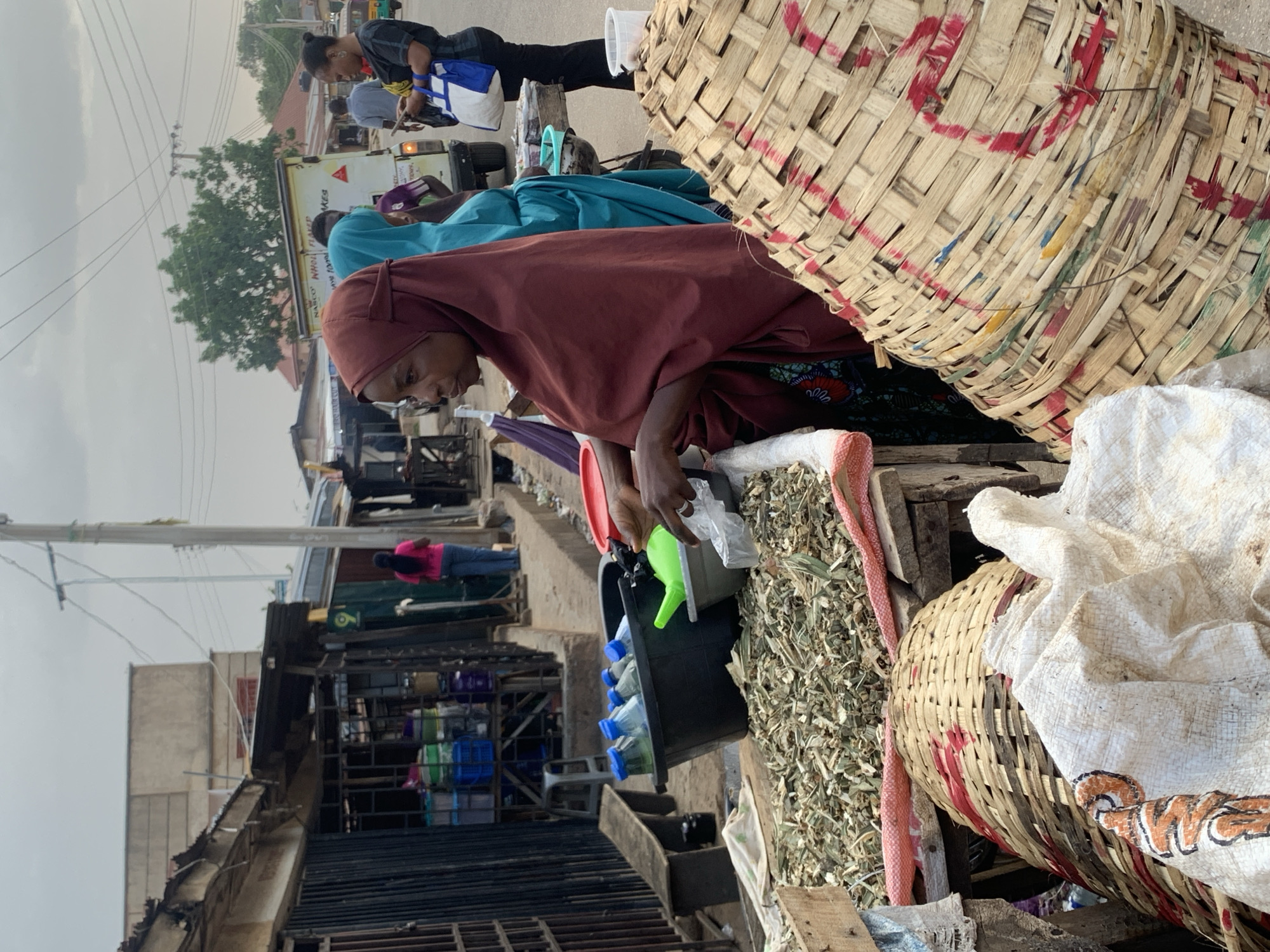 In Jos, Nigeria, a woman wearing a maroon headscarf and brown dress is at a market stand, putting small plastic bags in a black container with a green funnel. There is a pile of what appear to be dried herbs or leaves on the wooden table. Baskets made of woven material are to the right of the woman. Bottles are visible in the container. Other people are in the background, along with a light-colored truck. Buildings and electrical wires are in the background. The day appears overcast.
