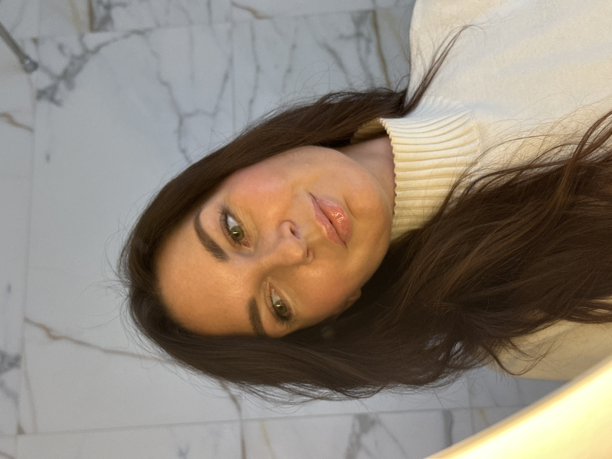 A woman with long, dark brown hair and fair skin is wearing a cream-colored turtleneck sweater. She has green eyes and glossy lips. The background appears to be a bathroom with marble or tile walls. The location is not specified in the image, nor can the location of Chaikovsky, Russia be determined.