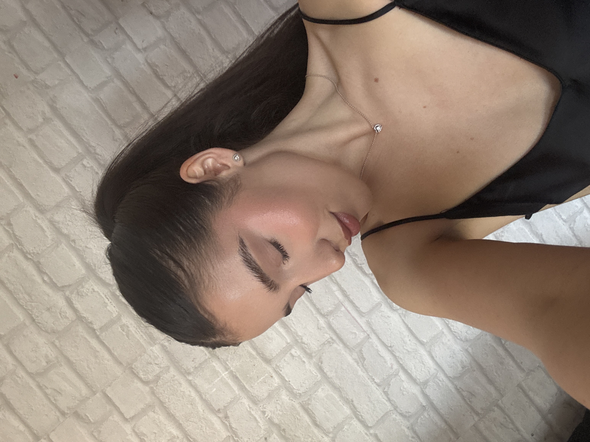 A young woman is shown in a close-up shot. She has dark hair and is wearing a black top. A silver necklace with a small pendant is visible around her neck. She has pierced ears and is wearing earrings. The background appears to be a white brick wall. The location and city are unknown.