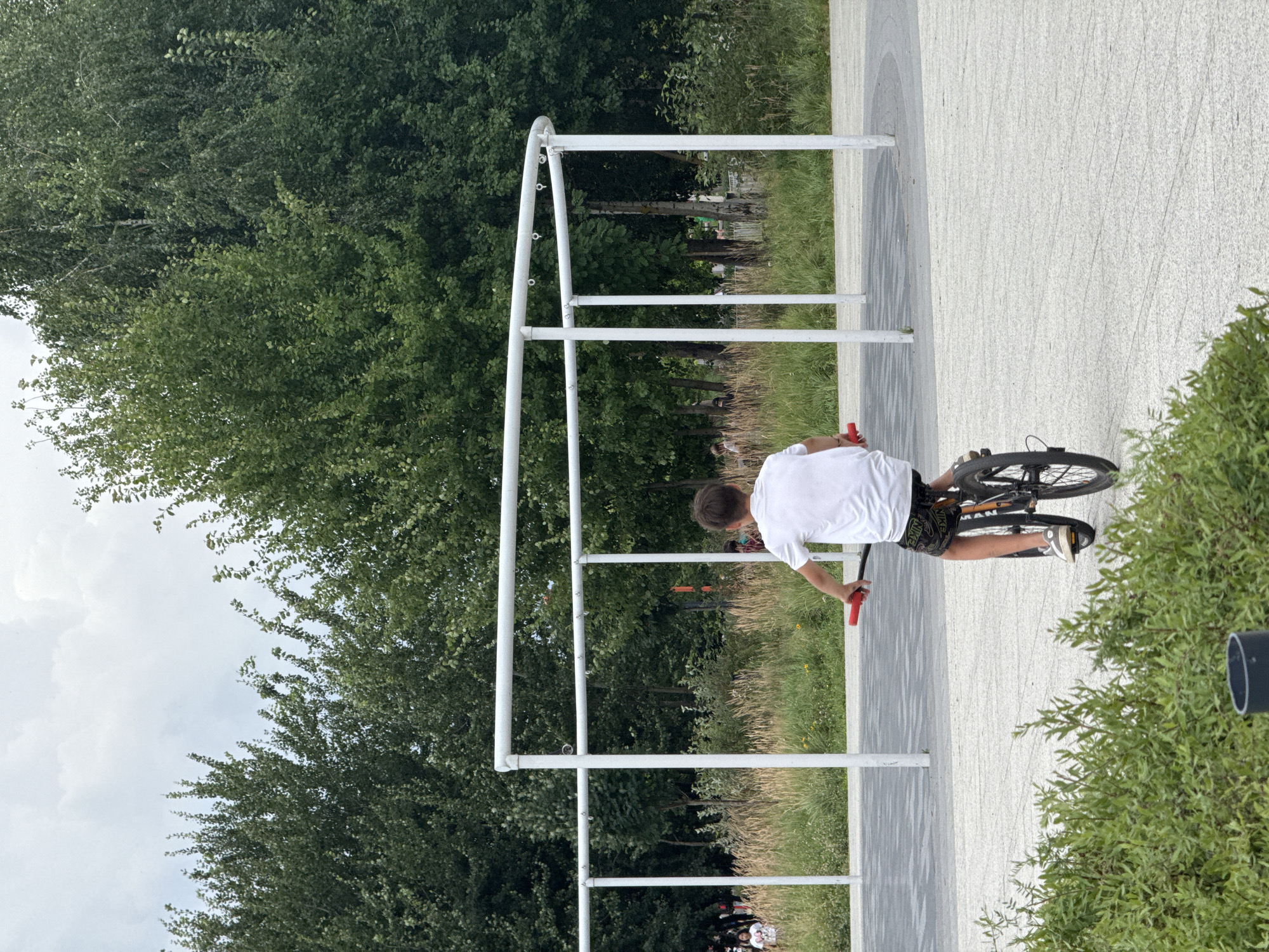 The image depicts a boy riding a bicycle on a paved area surrounded by greenery and trees. The boy is wearing a white t-shirt and patterned shorts, and is seen from behind. In the background, there is a modern architectural structure with white poles and curved bars. Other people can be seen in the distance. The sky is overcast. The location is the "URBICA" park in Kazan, Russia.