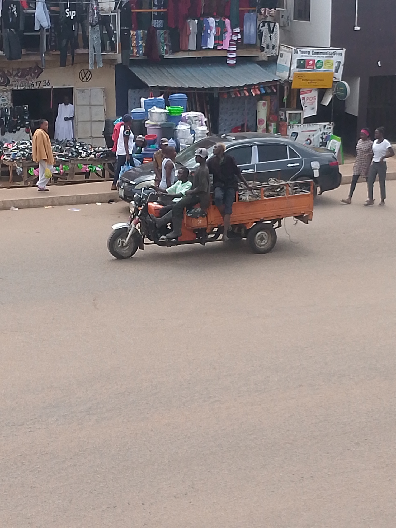 The image depicts a street scene in Bukuru, Nigeria. An orange motorized tricycle, carrying three people and a load of what appears to be fish or dried goods, is moving on the paved road. The driver is wearing a light green shirt, the second passenger is wearing a dark gray shirt, and the third is wearing a dark shirt and denim shorts.
Behind the tricycle, a dark-colored sedan is visible. Two women are walking on the sidewalk nearby. One is wearing a patterned dress, and the other is wearing a white shirt and gray pants. On the left side of the image, a store with clothes and shoes displayed outside is visible. Across the street, a sign for "Da Young Communications" is displayed. The environment is a bustling urban setting.