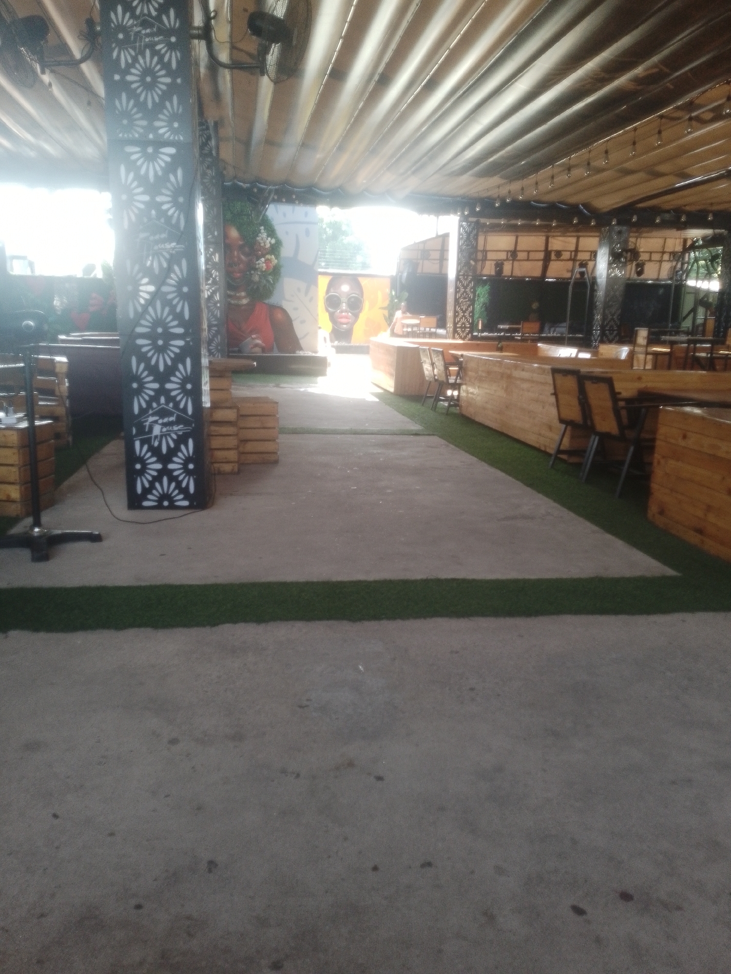 The image depicts an outdoor seating area at a venue identified by the text "Vibes House" on a decorated pillar. The seating is arranged around wooden tables and chairs, with some tables featuring built-in seating. The area is covered by a corrugated roof structure, supported by pillars and beams, with artificial lighting visible. The ground surface is a mixture of concrete and artificial turf, with the turf forming a green border. In the background, murals of people are visible on the walls, adding to the ambiance of the venue. The overall scene suggests a relaxed and social environment, likely a restaurant or bar, possibly located in Howayi, Nigeria.