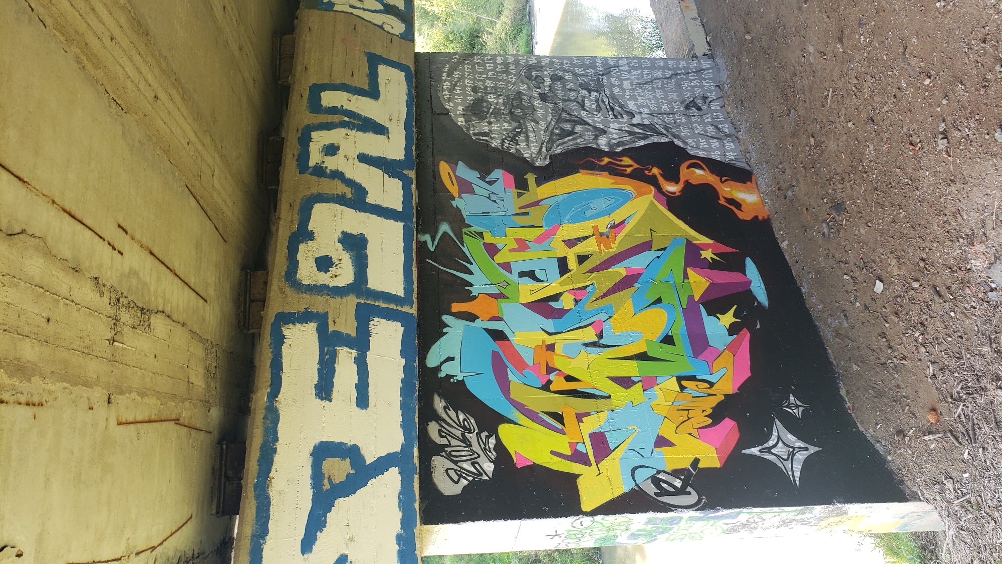 The image depicts a section of an underpass in Prešov, Slovakia, featuring prominent graffiti art. The ceiling of the underpass, made of concrete, shows signs of wear and tear, with exposed rebar. A large, horizontal concrete beam, painted white with thick blue outlines of letters, runs across the scene. Below this beam, the concrete wall is covered with vibrant graffiti. A central piece of graffiti is a complex, colorful arrangement of abstract shapes and letters in shades of yellow, green, blue, pink, and orange. To the left of this main piece, the number "2016" is written in white with a black outline, and some stylized star shapes are visible lower down. To the right of the colorful graffiti, a monochromatic depiction of a face or head is rendered, seemingly composed of densely packed numbers and text. Flames are painted on the wall between the colorful graffiti and the numbered face. The ground beneath the underpass is covered with dirt, small stones, and debris. In the background, a body of water, likely a river or stream, is visible, with trees and foliage on the opposite bank, suggesting an outdoor, possibly semi-urban or park-like setting. The lighting suggests daytime.