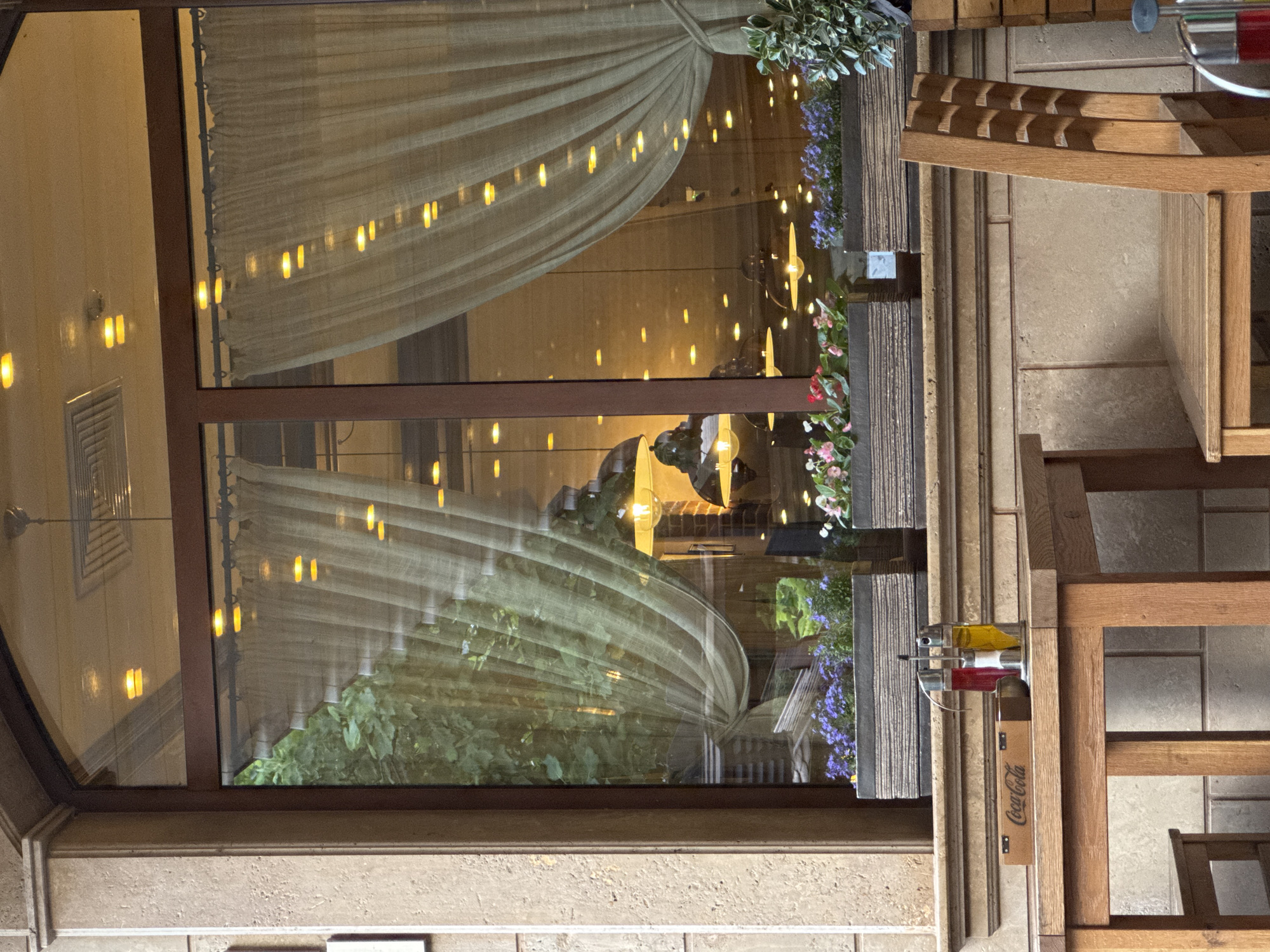 The image shows an outdoor seating area of a restaurant or cafe with a large window. The window reflects interior lights and greenery. On a wooden table in the foreground, there is a Coca-Cola branded box, a salt and pepper shaker set, and a bottle of olive oil or dressing. In front of the window, there are two planter boxes filled with flowers, one with purple flowers and the other with pink and white flowers. To the right of the table, a wooden chair is visible. The scene is observed from a low angle, with the focus on the table and chair.
