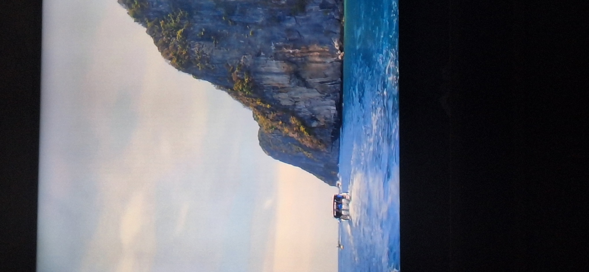 The image depicts a body of water with a large, rocky island covered in greenery on the right side. Several boats are visible on the water. One boat in the foreground appears to be a tour boat with people on it, moving away from the viewer. The sky is light blue with some clouds. The scene is likely a tropical or coastal environment. There is no information in the image that would indicate the location is in the city of Bila Tserkva, Ukraine.