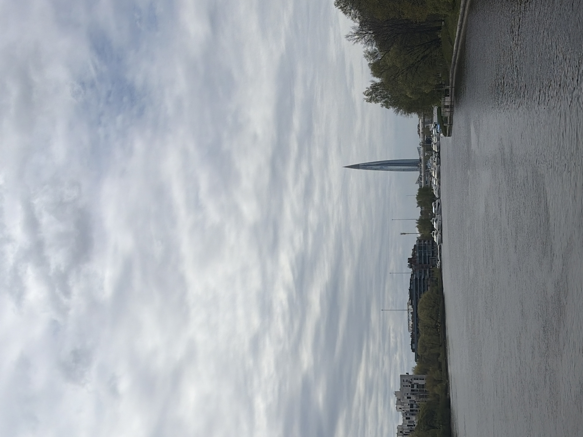 The image shows a cityscape of Saint Petersburg, Russia, seen from a waterfront perspective. The dominant feature is the Lakhta Center skyscraper, recognizable by its spire-like design. A body of water runs alongside the city, and several boats are docked along the shore. Buildings of varying heights line the waterfront, and trees are scattered among the structures. The sky is overcast with textured clouds.