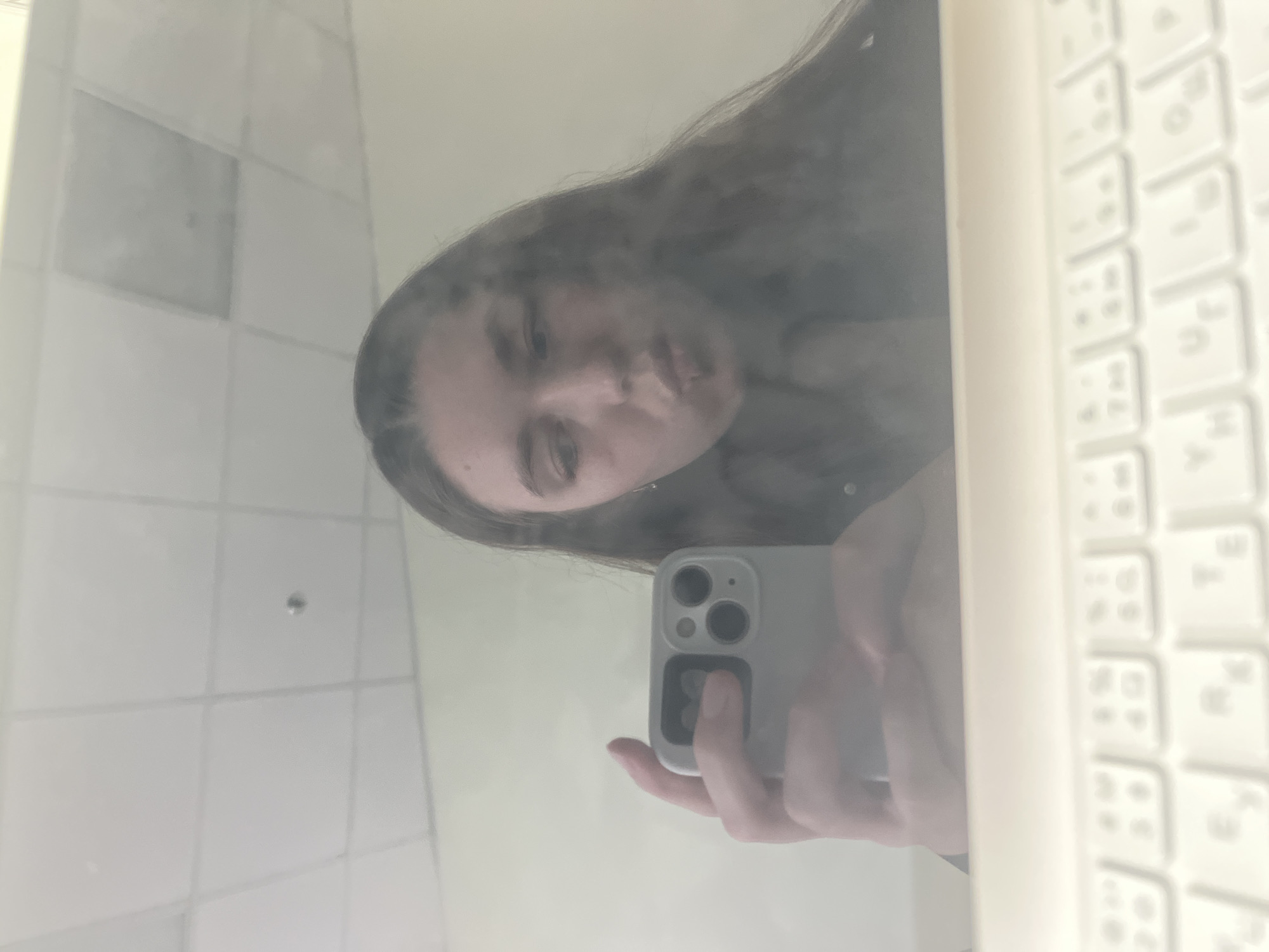 A person with long brown hair is holding a gray smartphone in their left hand, taking a selfie in the reflection of a white laptop screen. The reflection reveals a bathroom with white tiled walls. The location is potentially a hotel room or residence in Moscow, Russia, though no specific landmarks are visible to confirm this. The person's face is partially obscured by the phone. The laptop keyboard is visible on the right edge of the frame.
