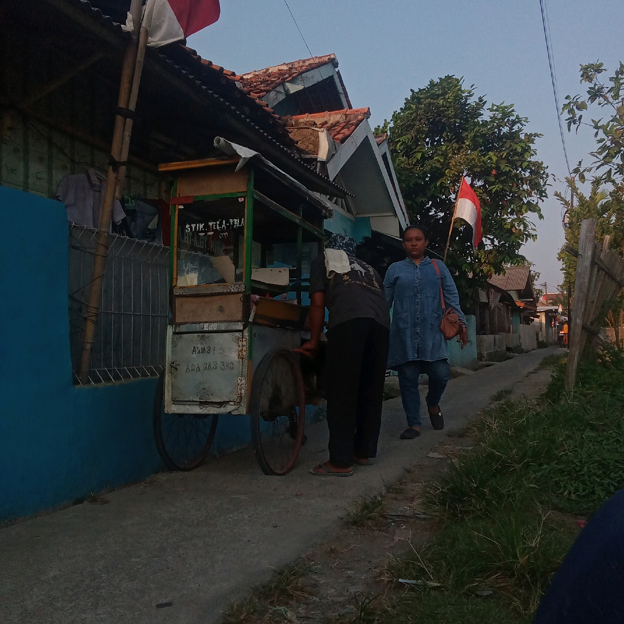 The image depicts a street scene in Karawang, Indonesia, likely during the late afternoon or early evening due to the soft lighting. In the foreground, a woman in a blue denim dress and jeans walks away from the camera, carrying a brown purse. To her left, a man wearing a dark t-shirt and dark pants is seen from behind, seemingly engaged with a food cart. The food cart itself is colorful, with green and yellow accents, and displays various items, including what appears to be a sign with Indonesian text, "STIK. TELA-TELA." Another sign on the cart reads, "AWAS! ADA GAS 3KG." The cart has two wheels and is positioned on a paved walkway that runs alongside a vibrant blue wall. Beyond the cart, a modest house with a tiled roof is visible. A red and white Indonesian flag is planted near the house. The surrounding environment includes trees and vegetation along the side of the walkway, with a wooden fence on the right. The overall impression is of a residential street with a small business operating from a mobile cart.