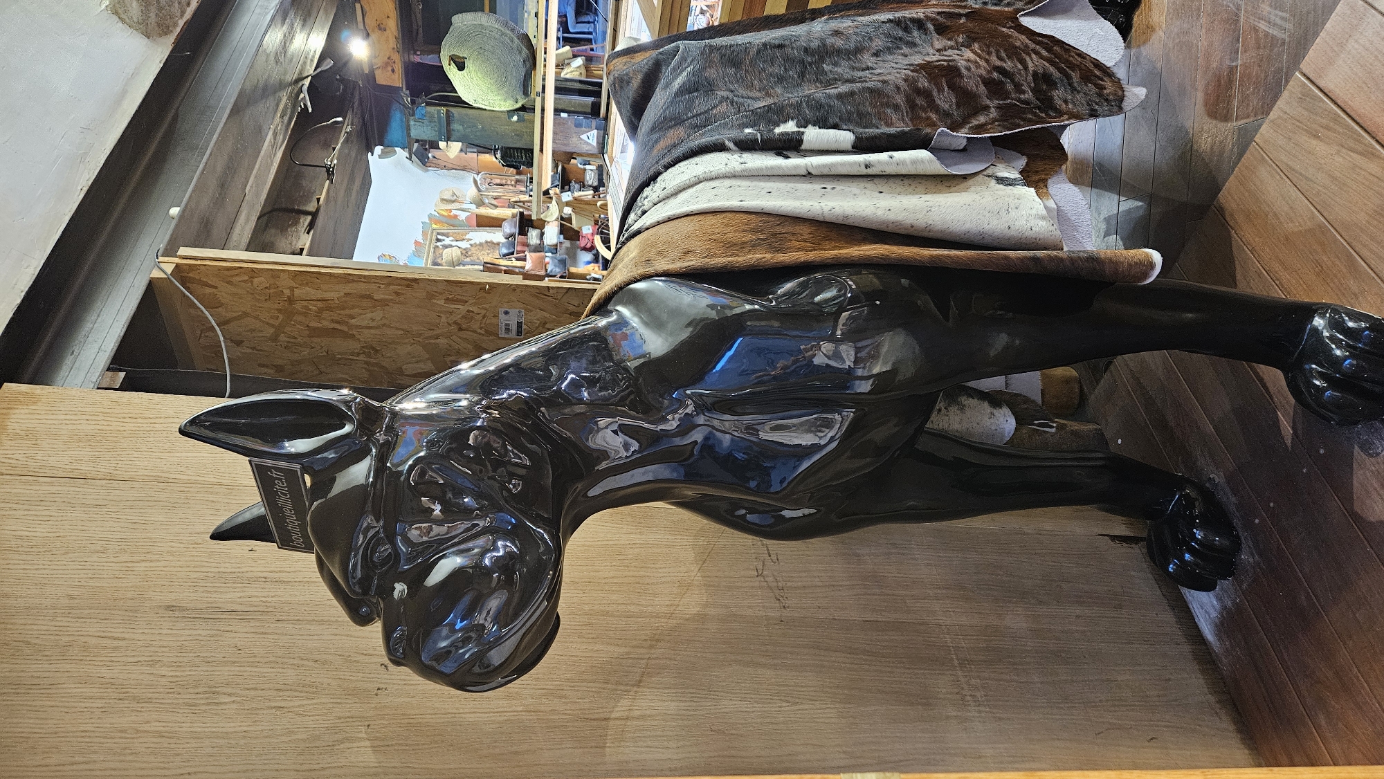 A life-sized, glossy black statue of a dog, possibly a boxer breed, stands in a retail environment. A sign reading "boutiquillicite.fr" is affixed to the statue's forehead. The dog statue is draped with several hides or pelts of varying patterns and colors, including brown, white, and spotted. Behind the statue, a wooden wall panel and shelves stocked with merchandise are visible. The floor is made of polished wooden planks. The background shows a section of a shop interior, with more shelves and objects, suggesting it might be a boutique. The overall lighting is warm and ambient, with some reflections visible on the statue's surface. The scene appears to be located in the city of L'Île-Rousse, France, likely within a shop specializing in animal-themed decor or animal product retail.
