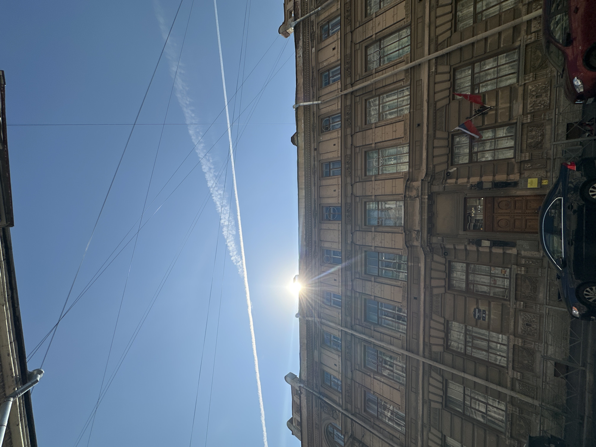 The image shows the architecture and urban landscape of Saint Petersburg, Russia. An old building with multiple windows and architectural details is visible. The sun is directly behind the building, creating a bright glare. Above the building, there is a clear blue sky with vapor trails. Power lines are also strung across the sky. A dark blue vehicle is visible on the right side of the building and a red vehicle is also visible at the top right. Two sets of red and white striped flags are fixed between windows on the facade. A sign that reads "Улица Союза Печатников, 26" is visible on the facade.