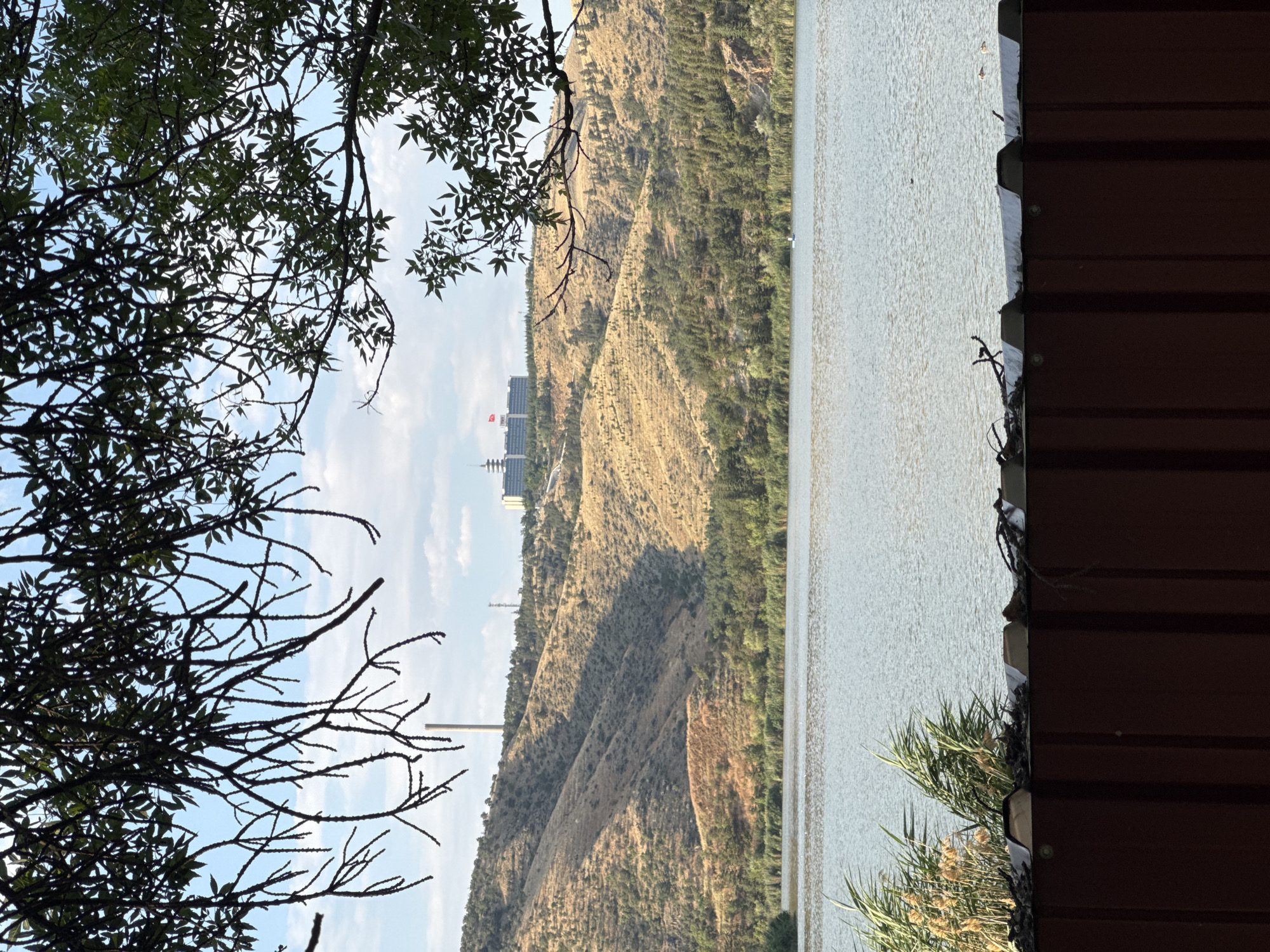 The image captures a serene view of a lake in Gölbaşı, Turkey, framed by the silhouette of tree branches in the foreground. Beyond the water, rolling hills dotted with vegetation lead up to a distant cityscape. A prominent feature on the hill is a tall, white chimney, likely part of an industrial or utility structure, accompanied by a modern building complex on the crest. The water itself is calm, reflecting the sky and surrounding landscape, with a few scattered reeds along the bank. The overall atmosphere is peaceful, hinting at a natural landscape within an urban or semi-urban environment.