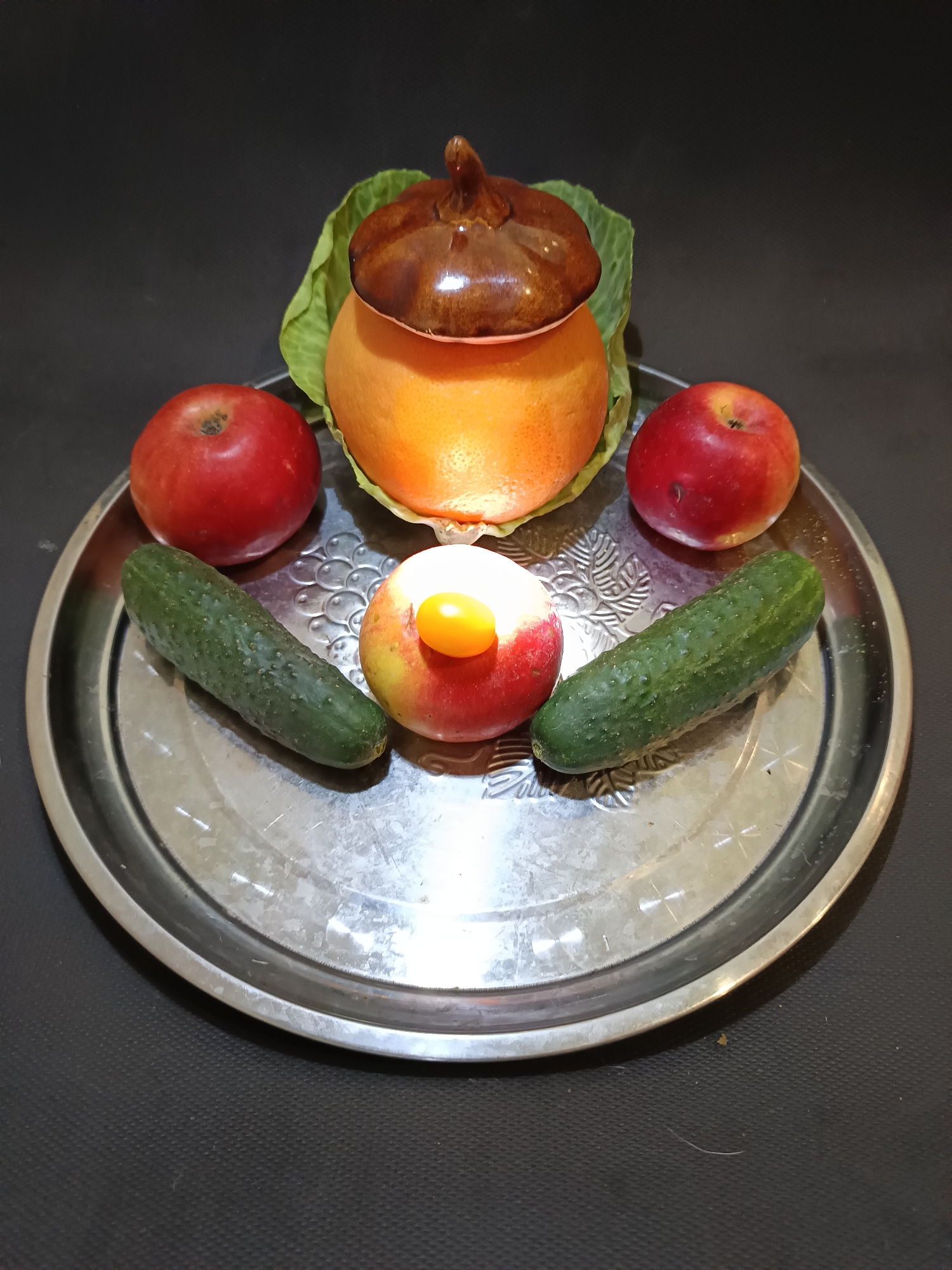 A still life arrangement of fruits and vegetables is presented on a round, metallic tray. The tray is centered in the frame and has a decorative, embossed pattern around its rim.
Dominating the center of the arrangement is a large, round orange, partially covered by a brown, ceramic lid shaped like a pumpkin. Beneath the orange, a large green leaf, likely from a cabbage, serves as a base.
Surrounding the central element are two red apples, one on the left and one on the right. Between the apples and closer to the front of the tray are two green cucumbers. In the very center of the arrangement, on top of one of the apples, is a small, bright yellow object, possibly a cherry tomato or a small pepper.
The background is a dark, solid color, likely black or a very dark grey, creating a stark contrast with the colorful fruits and vegetables. The lighting appears to be artificial and somewhat direct, casting subtle shadows on the tray and its contents.
No people are visible in the image, and no specific activity or event is taking place. The overall impression is one of a simple, almost artistic, arrangement of natural produce. There is no visible text in the image. The time of day, weather, and specific location are not discernible from the image.