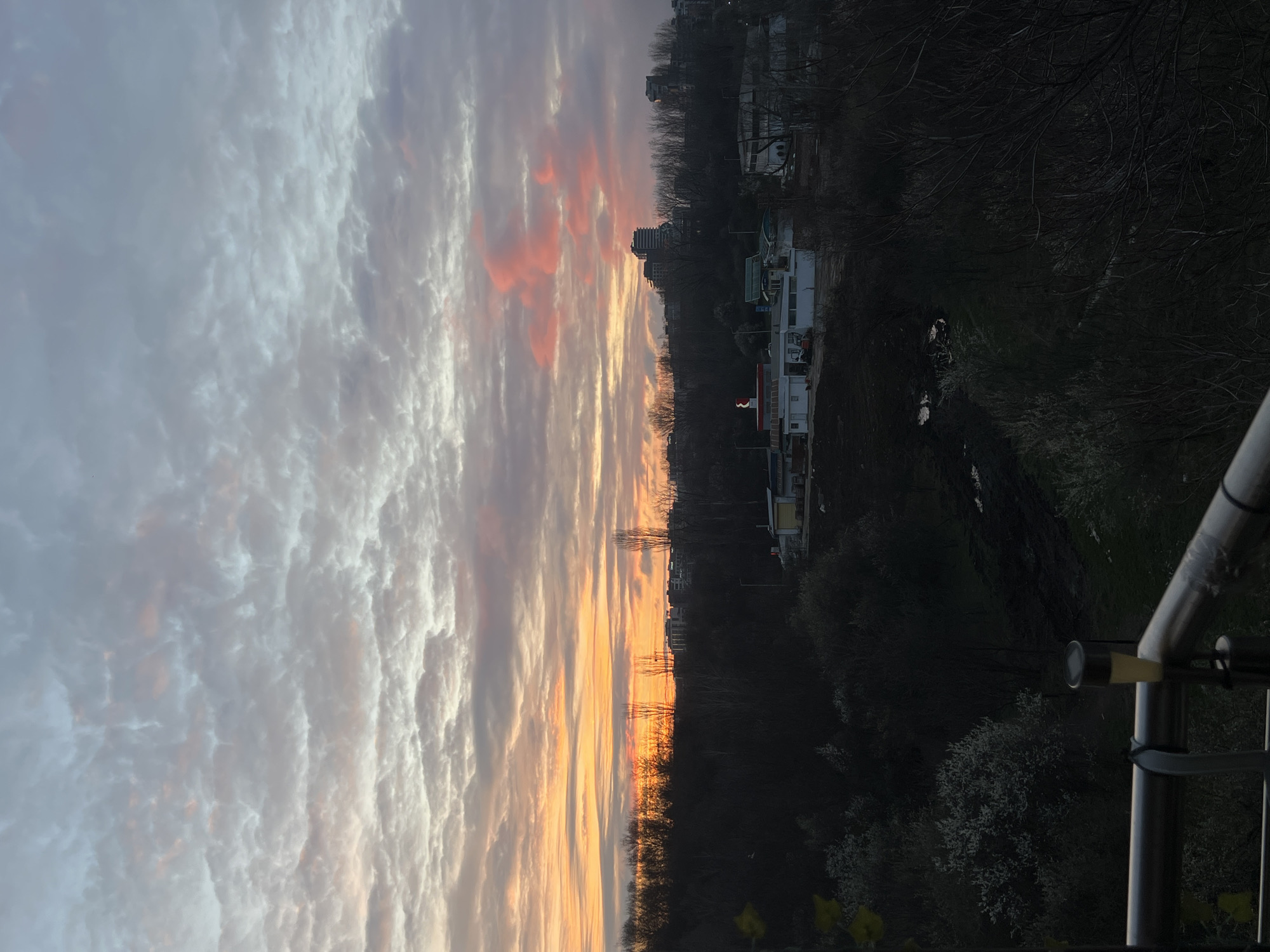 The image captures a sunset scene, likely taken from a balcony overlooking a cityscape in Sofia, Bulgaria. The sky is dominated by a layered cloud formation, transitioning from white to pink to orange hues towards the horizon. The buildings and infrastructure are dimly visible amidst dark trees and vegetation. A section of a metal balcony railing is visible in the foreground.