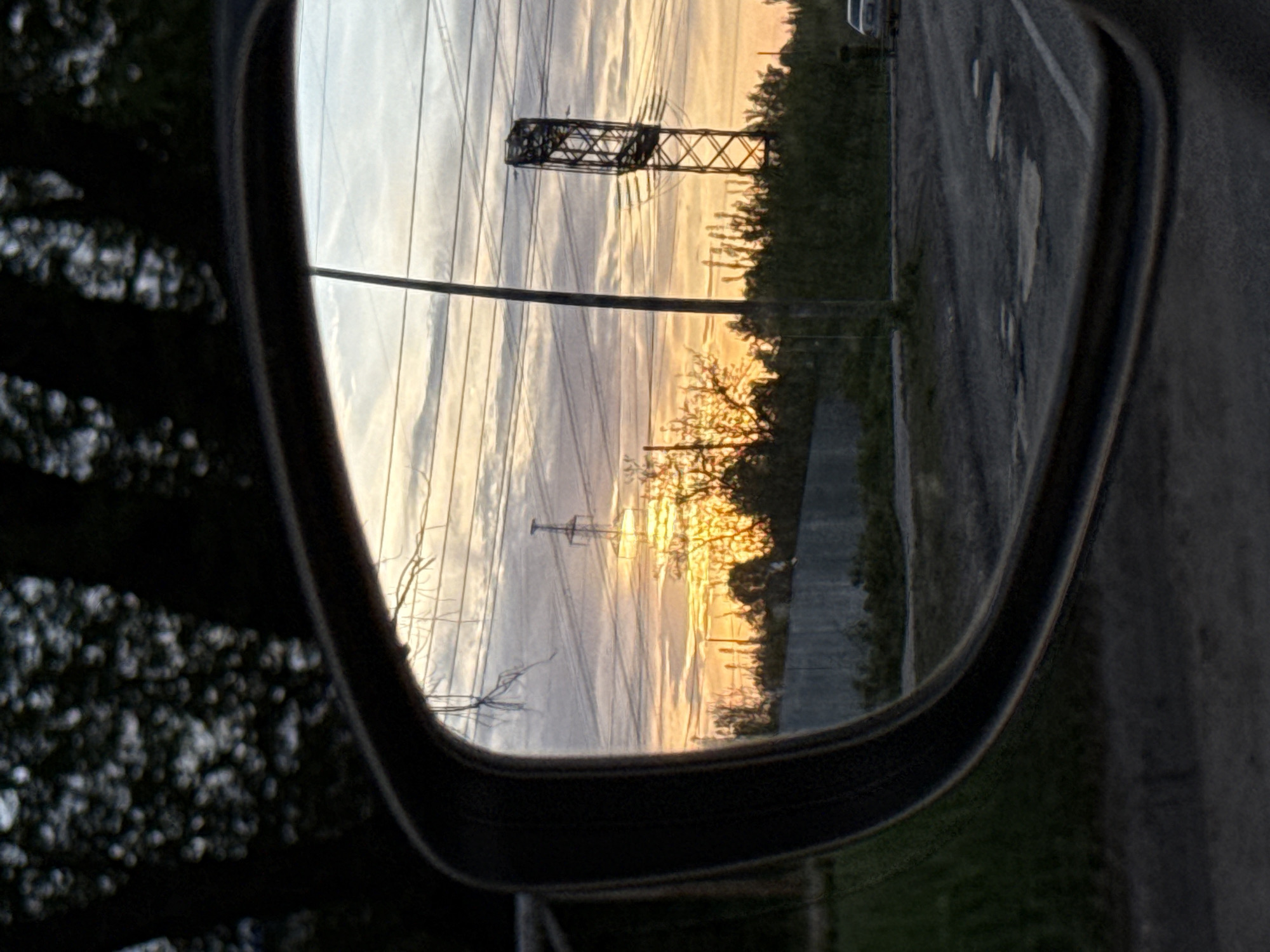 The image shows a view of a sunset reflected in a car's side mirror. Power lines and towers are visible in the reflection, along with some trees and a road. The car's mirror frame is black. The location is speculated as being in Chaikovsky, Russia.