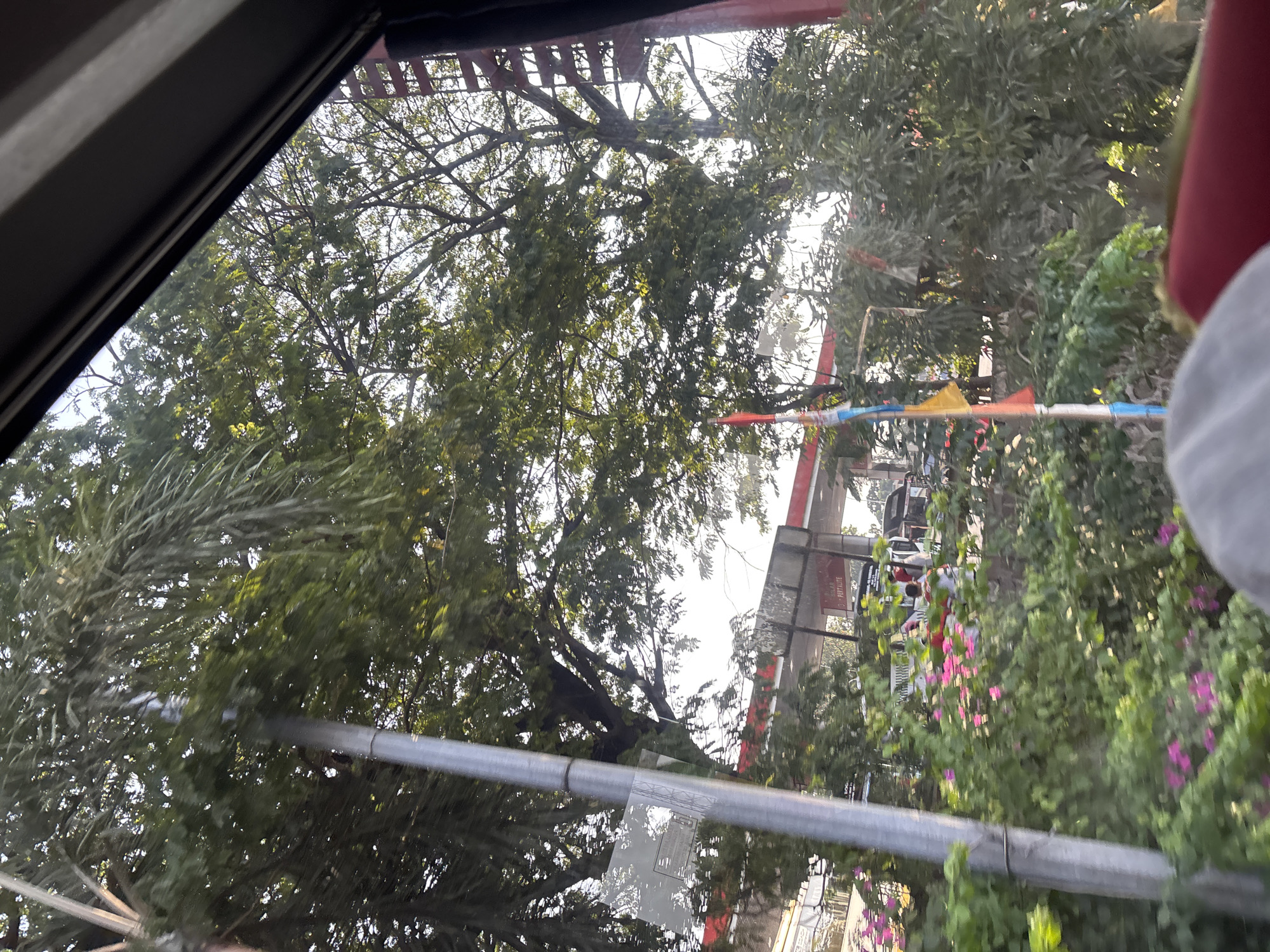 The image captures a view from inside a vehicle, looking out at a gas station and its surroundings. A tall, grey pole is visible on the left side of the frame, positioned in front of lush green foliage and trees. In the background, a gas station canopy with red accents and signage is present. Several colorful flags are strung up, adding a festive touch. The scene suggests an outdoor environment in a city, likely during daylight hours, with the light filtering through the trees. The presence of a gas station and the glimpse of vehicles and people indicates a public commercial area. While not explicitly stated, the foliage and trees suggest a tropical or subtropical climate, common in Indonesia.