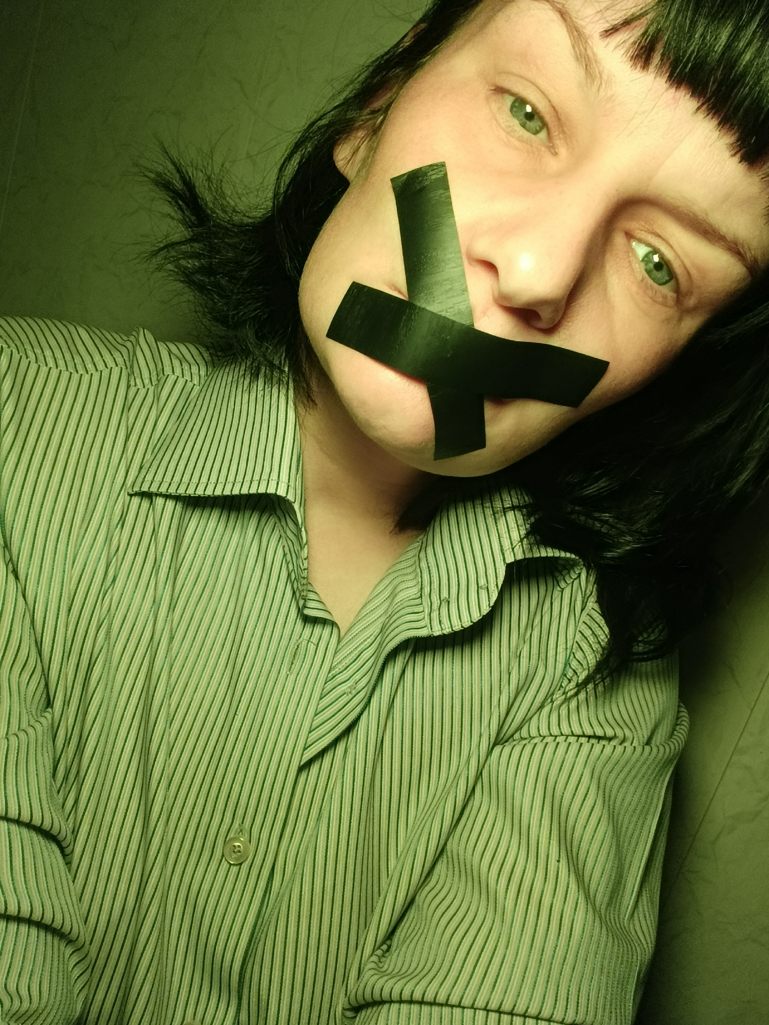 A close-up shot shows a person with light skin and short, dark hair, including bangs. The person is wearing a green and white striped button-down shirt. Black tape is placed in an "X" shape over their mouth. The person is looking slightly downward and to the side. The background appears to be a solid, light-colored wall. No evidence suggests that the image was taken in the city of Фоки, Russia.