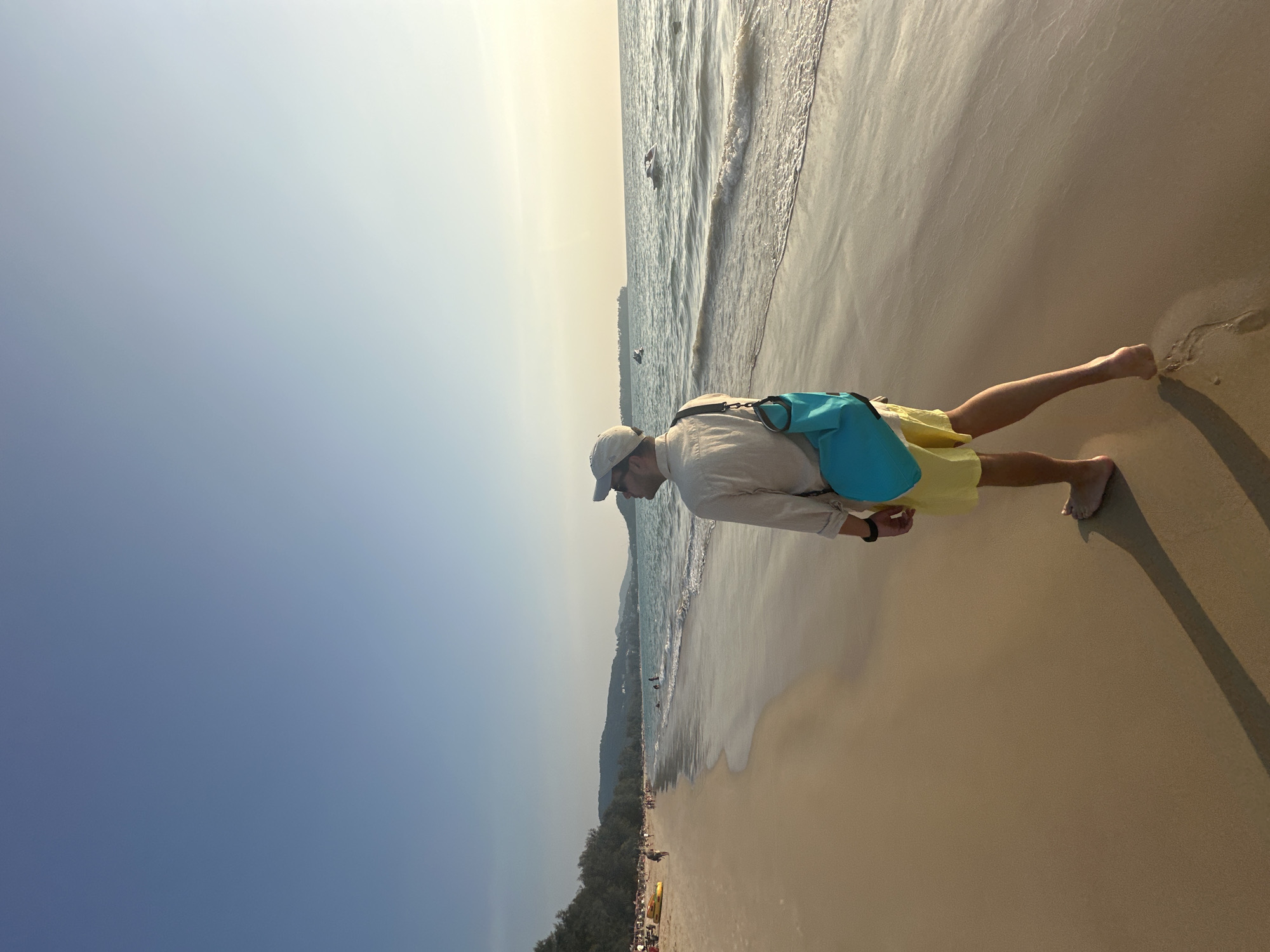 A barefoot individual walks along the wet sand of a beach in Choeng Thale, Thailand, under a clear sky during what appears to be late afternoon or early evening. The person, viewed from behind and slightly to the left, wears a light-colored long-sleeved shirt, yellow shorts, a light-colored baseball cap, and sunglasses, with a turquoise blue dry bag slung over their left shoulder. The individual casts a long shadow on the sand, extending towards the lower left of the frame, and is looking out towards the ocean on the right.
To the left, the beach extends into the distance, populated by other individuals, some seated under beach umbrellas, and several yellow kayaks or paddleboards are visible on the sand. A line of dense green foliage and trees borders the upper edge of the beach, backed by distant hills.
To the right, the ocean displays light waves breaking on the shore, with deeper water beyond. Several personal watercraft (jet skis) are visible on the water further out, and a low, distant landmass or island can be seen on the horizon. The sunlight appears to be low, casting a golden hue over the water and creating strong reflections on the wet sand.
