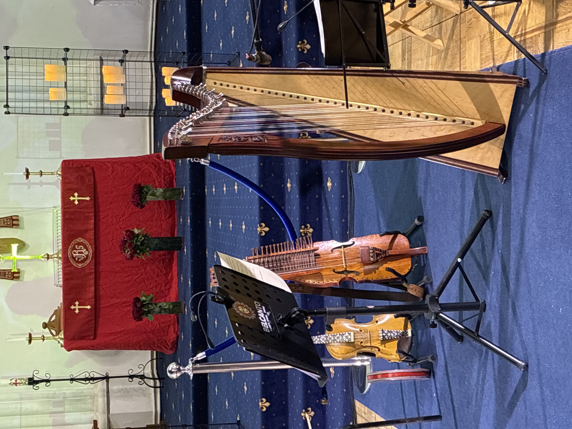 The image shows a room with musical instruments set up for performance. A harp is positioned prominently, next to a bowed string instrument, possibly a Hardanger fiddle, and a violin. A music stand with sheet music is also present. In the background, an altar draped with a red cloth featuring a cross and floral arrangements is visible. A metal shelf unit with artificial candles stands behind the altar. The room's carpet is blue with a fleur-de-lis pattern. The location is indeterminable, and there is no information that suggests it is in Moscow, Russia.