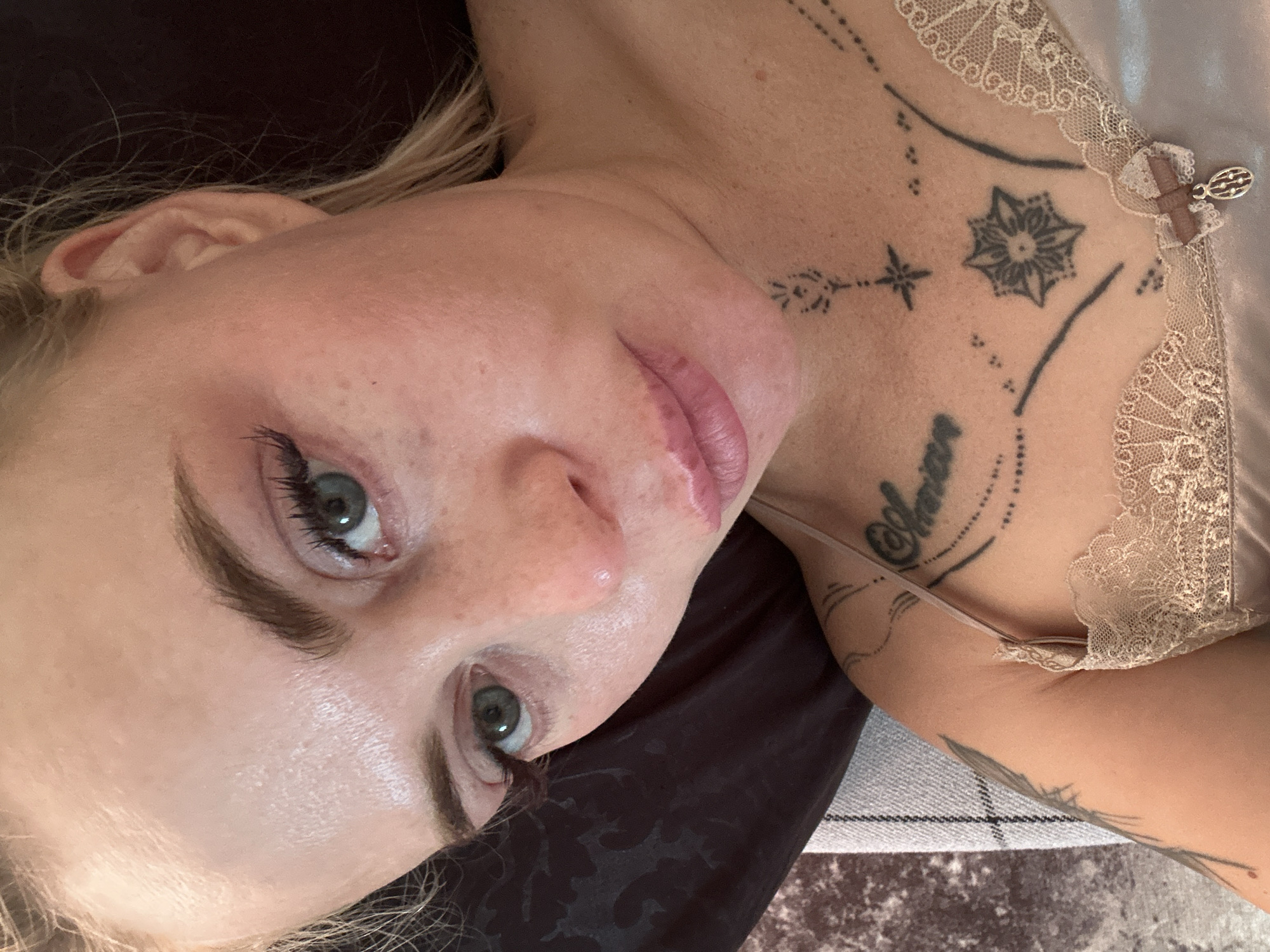 The image is a close-up, overhead shot of a woman's face and upper chest. She is lying down, with her head slightly tilted and her gaze directed upwards. Her blonde hair is visible around her face and shoulders. She has light skin with some freckles and what appear to be natural eyelashes. Her eyes are green, and her lips are slightly parted. A tattoo is visible on her chest. She is wearing a tan camisole with lace trim. The background is dark and appears to be a pillow or fabric. The scene is indoors, with no identifiable city or location details.