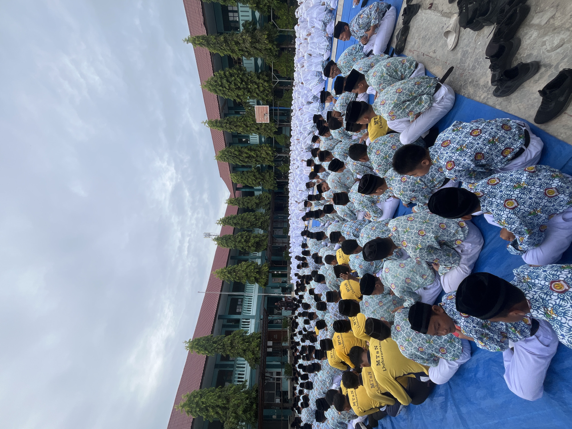 This image depicts a large group of students, predominantly young boys, sitting on a blue tarp in what appears to be a school courtyard in Lhokseumawe, Indonesia. The students are seated in rows facing a building with a red roof and green walls. Many of the boys are wearing white pants and patterned shirts with blue and white designs, along with black peci (caps). Some students in the foreground and middle ground are wearing yellow shirts with the text "MTsN Lhokseumawe" on them. The environment includes tall, slender green trees lining the building and a cloudy sky. In the background, a large group of students, possibly girls, are also visible, many wearing white headscarves. The overall scene suggests an outdoor assembly or event at an educational institution.