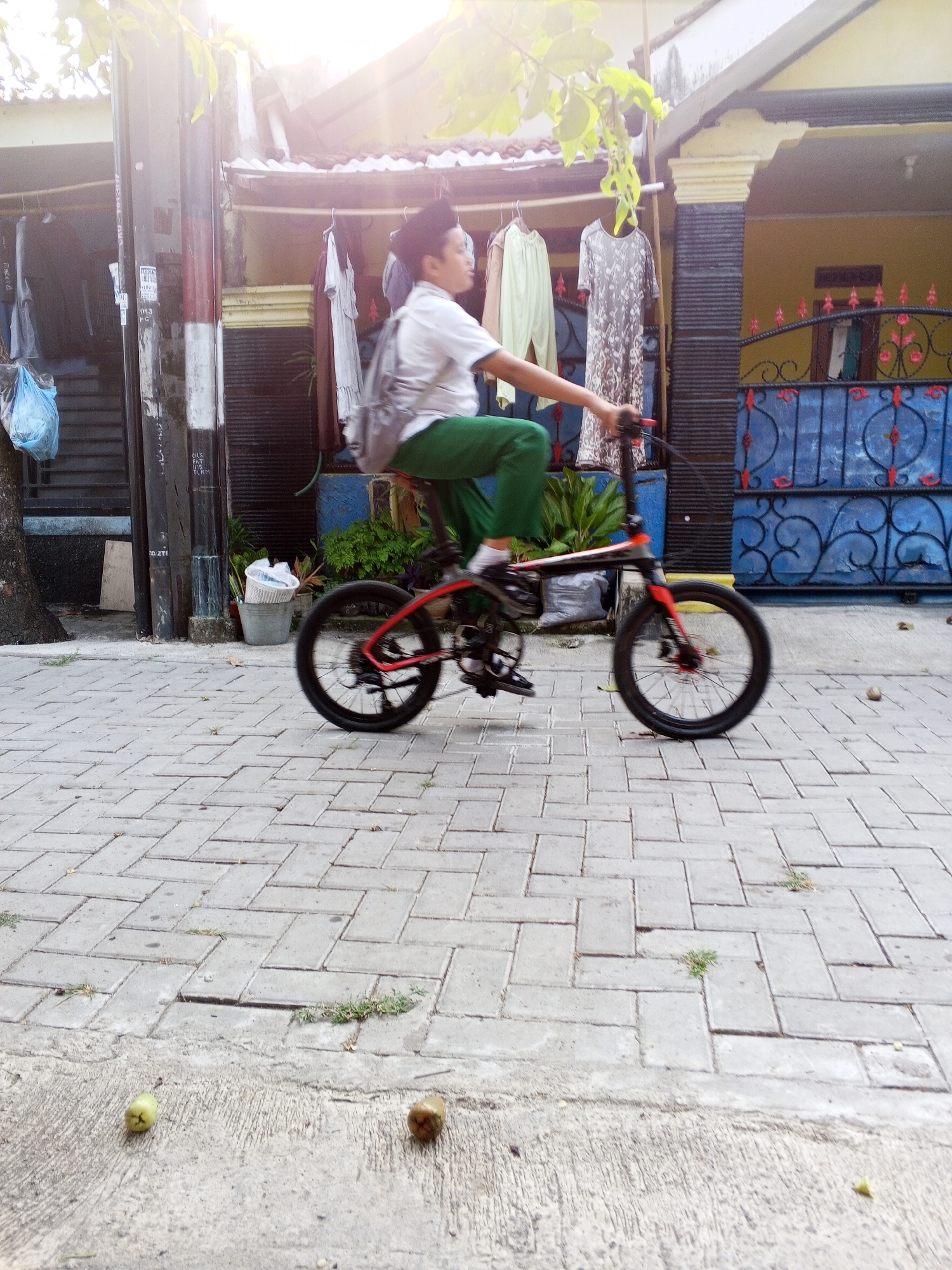 A young boy wearing a white shirt, green pants, and a backpack is riding a black and red bicycle on a brick-paved sidewalk. The boy is in motion, as indicated by the blur of the bicycle wheels. He is facing to the right of the frame and appears to be looking ahead. The background features a building with yellow walls and a blue metal fence. Clothes are hanging on a rack in front of the building. The scene appears to be outdoors on a sunny day. This location is in Ciruas, Indonesia.