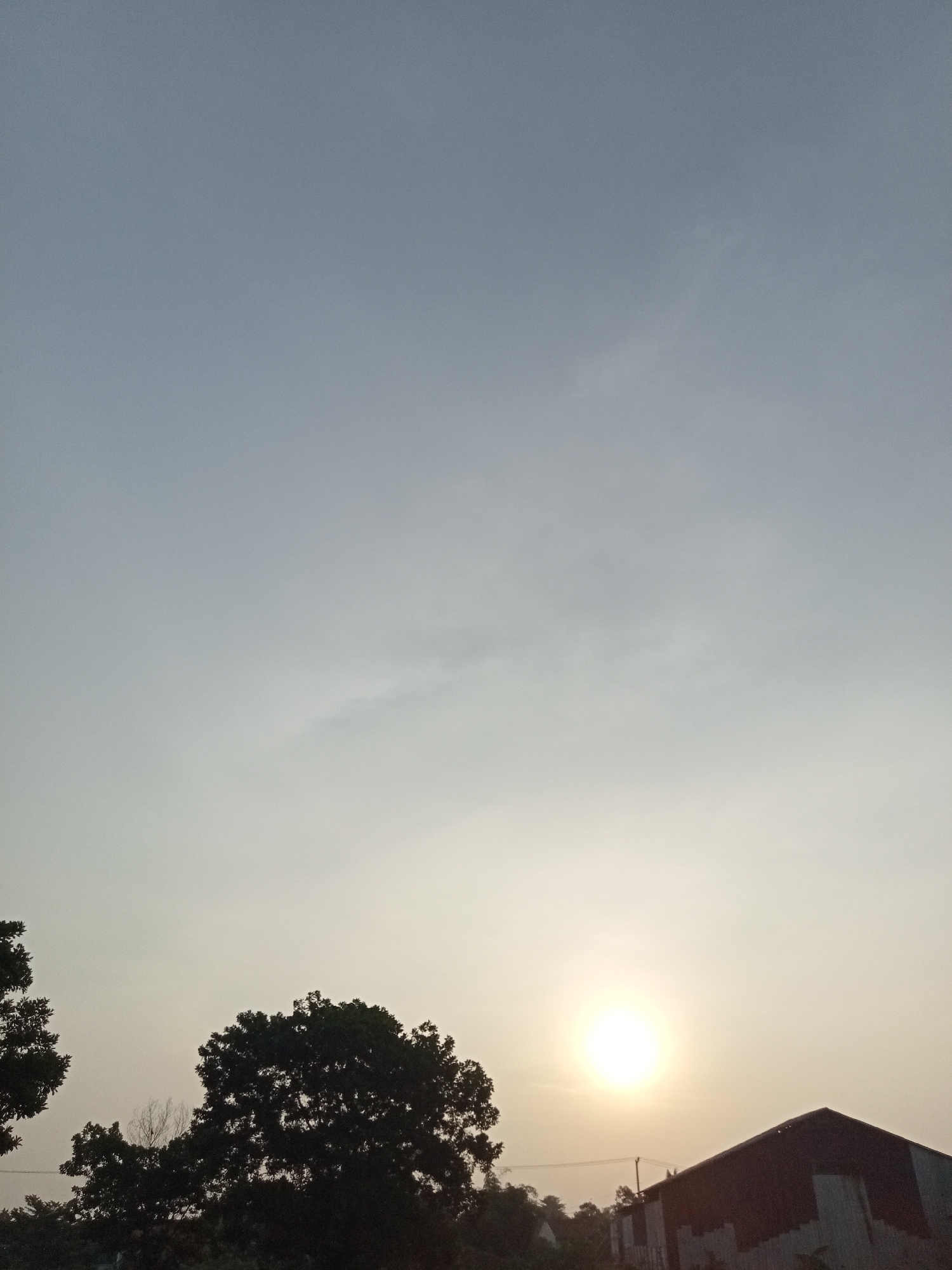 The image shows a silhouetted treeline and a building against a hazy sky with the sun visible as a bright orb. The treeline is dense with foliage, and a portion of a dark-roofed structure is visible on the right. The sky is a pale blue, gradually transitioning to a lighter hue near the horizon. The lighting suggests it is either sunrise or sunset. The environment appears to be outdoors, possibly in a semi-urban or rural area given the presence of the building and trees. There are no people or animals visible in the frame. The location is specified as Teluk Kenidai, Indonesia.