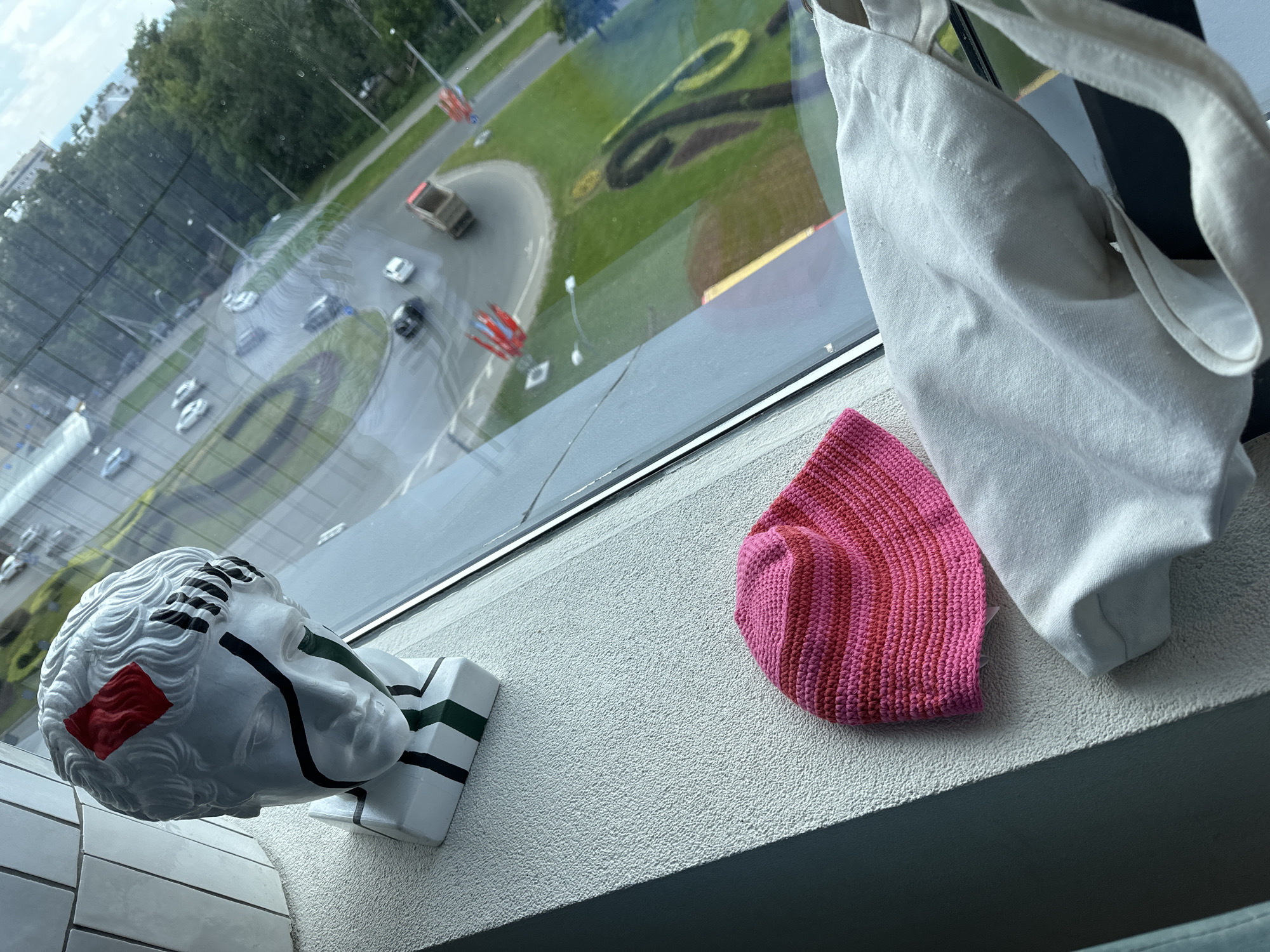 The image captures a scene from an indoor vantage point, looking out through a large window that overlooks an urban landscape in Nizhny Novgorod, Russia.
In the foreground, on a light-colored, textured windowsill, three main items are displayed. To the left is a white decorative bust, styled like a classical head but featuring modern, abstract embellishments: a bold red rectangular patch on the side of the head, black stripes on the wavy hair, a black line extending downwards from the left eye, and green and black stripes on its rectangular base. Adjacent to it, centrally placed on the sill, is a vibrant knitted item, possibly a hat or small bag, featuring alternating stripes of pink and a deeper red hue. To the right, a cream or white fabric tote bag hangs or rests near the window frame.
Through the window, an outdoor scene is visible, appearing under an overcast sky during daytime. A curved road with several cars, some blurred indicating motion, traverses a lush green area. Within this grassy expanse, large, stylized shapes, possibly decorative numbers or letters (one resembles a '2' or an 'S'), are formed into the lawn. Trees and distant buildings complete the urban background. Reflections on the window surface add depth and slight distortion to the external view. There is no discernible text within the image apart from the stylized shapes in the grass.