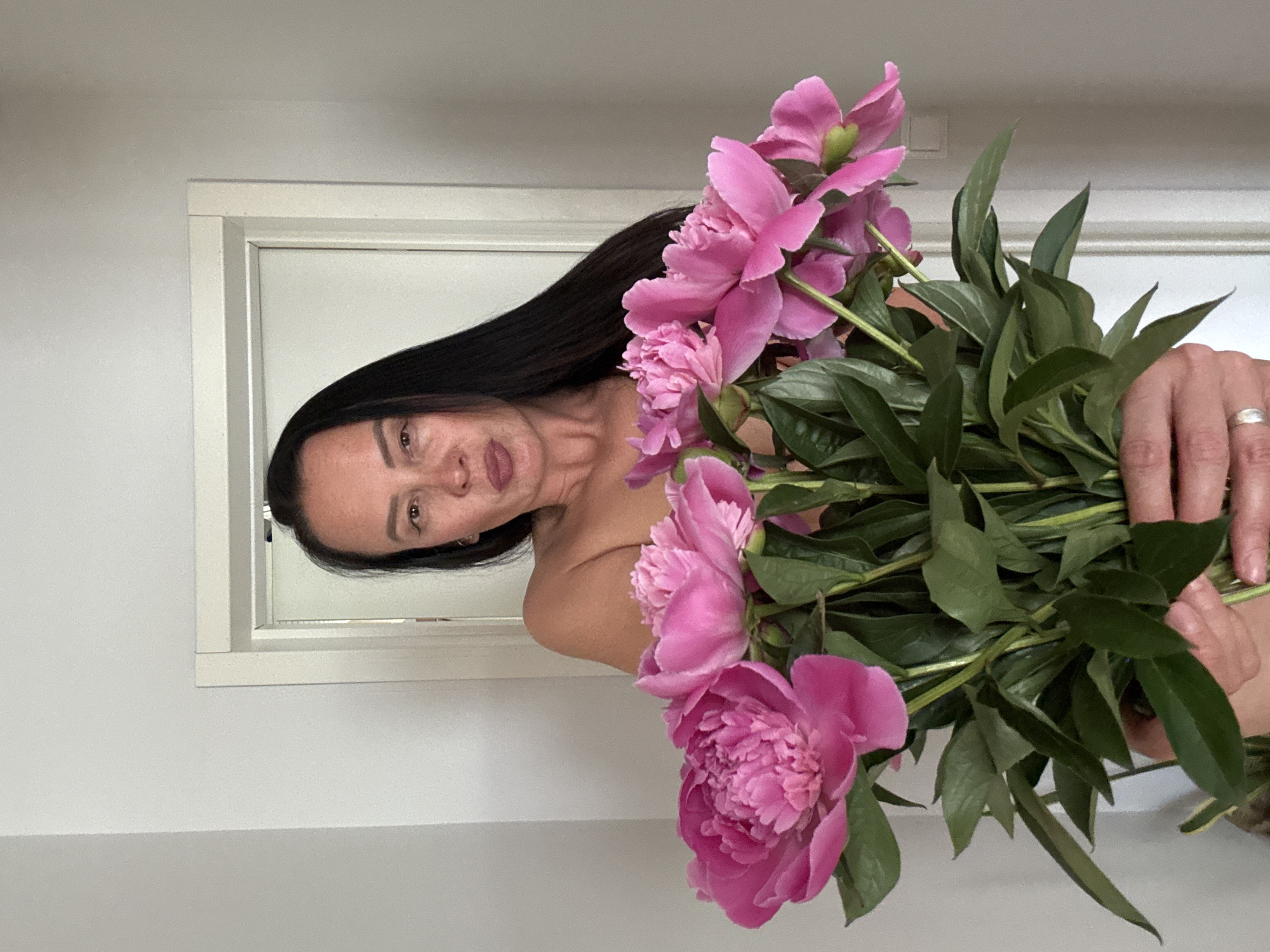 A woman with long, dark hair holds a bouquet of pink peonies in front of a white window frame. She is facing the camera with her head tilted. The background consists of a plain white wall. The image does not contain sufficient information to determine the location as Chaikovsky, Russia.