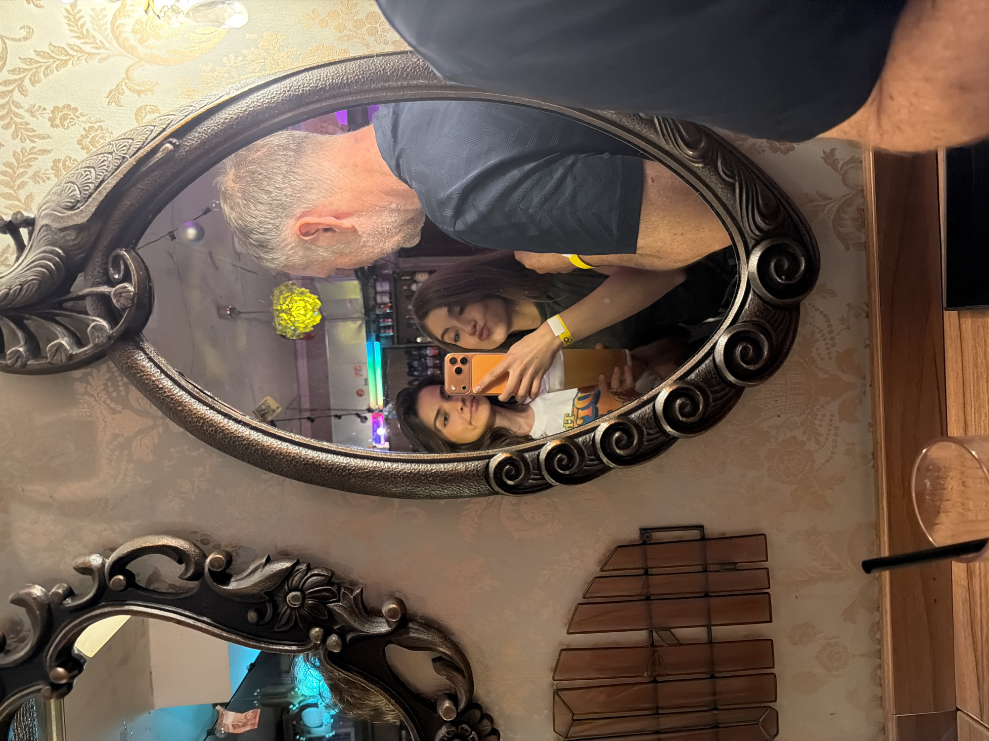 An ornate, dark-framed oval mirror is the central focus of this image, positioned against a light-colored wall with a gold patterned wallpaper. The original image is rotated 90 degrees counter-clockwise.
Reflected in the upper part of the main mirror is a man with gray hair, wearing a dark blue or black collared shirt with a textured pattern. Below his reflection, two women are visible in the mirror. One woman, with dark hair, is holding an orange-brown smartphone up to take a mirror selfie, making a slight pout with her lips. She has a yellow wristband on her right wrist. Next to her, another woman is reflected, looking towards the phone with a faint smile. Behind them, in the reflection, are colorful artificial lights, including a bright yellow decorative light and string lights, suggesting an indoor evening or night setting.
To the lower left of the main mirror, a portion of a second, similarly ornate mirror with a dark frame is visible, reflecting a bright, possibly artificial, light source. To the right of the main mirror, a wooden structure, possibly a cabinet or shelving unit, and part of a clear glass or cup can be seen.
The scene appears to be indoors, possibly in a establishment like a bar or restaurant, given the decorative elements, artificial lighting, and the presence of multiple individuals. The overall atmosphere seems relaxed and social, with the selfie-taking activity indicating a casual moment. No clear legible text is visible in the image. The location context provided is Moscow, Russia.
