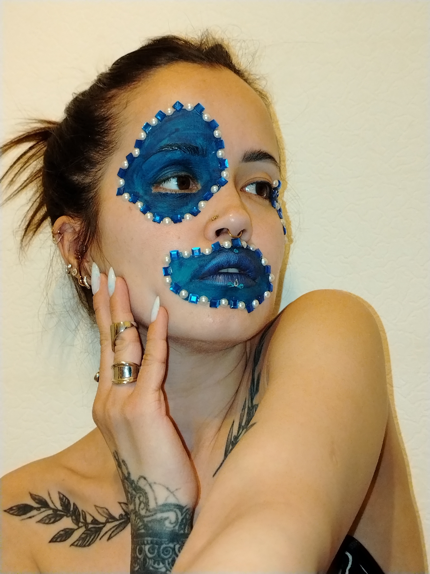 A person is posing in front of a pale, textured wall. The person's hair is pulled back into a loose bun, and they are adorned with blue face paint and small, white beads around the left eye and mouth. The lips are also painted blue. The person has several piercings in the left ear and a nose ring. The left hand is raised to the face, displaying long, white fingernails and several rings. A tattoo of a leafy branch is visible on the left shoulder, and another tattoo is on the left wrist. The person is wearing a black top. The background appears to be a plain wall. There is no indication of the location being Noviy, Russia.