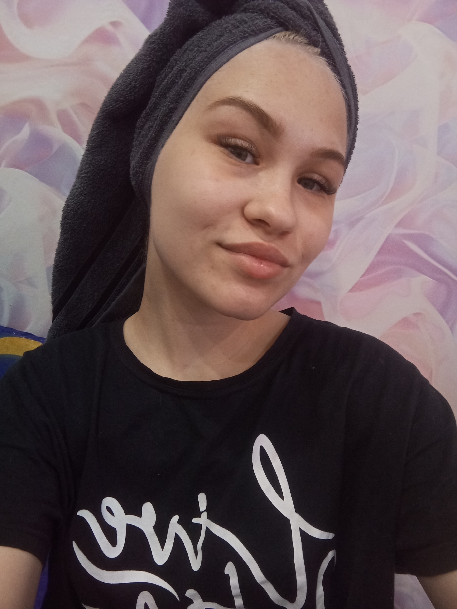 A woman is centered in the frame, posing in a selfie. She wears a black t-shirt with white lettering on the front and has a gray towel wrapped around her head. The background consists of a wall with a purple and white swirled pattern. The picture may have been taken in a personal residence in the city of Krasnodar, Russia, based on user query.
