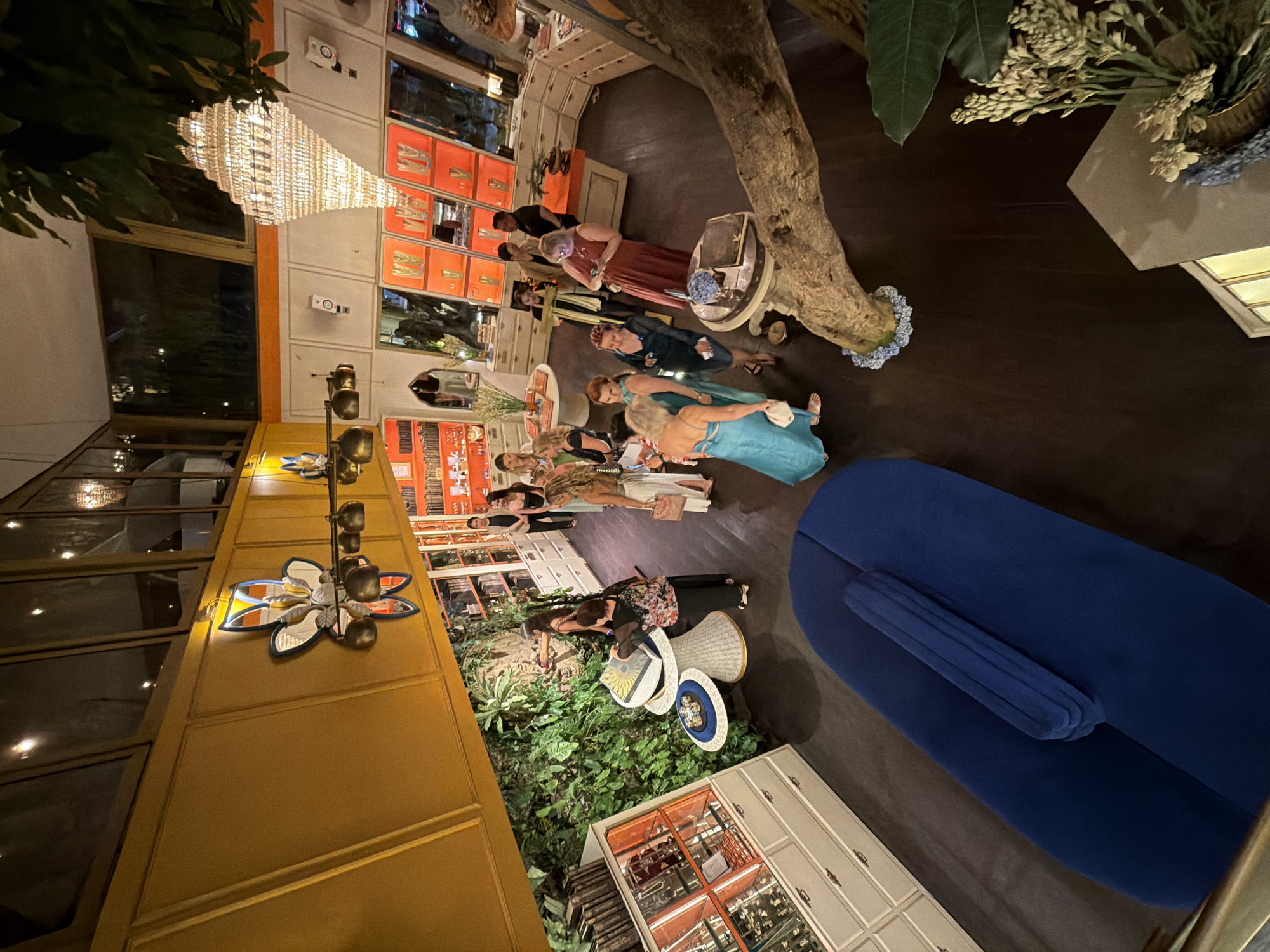 This is a high-angle, indoor, wide shot of a retail space, likely a boutique or gallery, with a tropical theme and vibrant decor. The room features a yellow upper wall and ceiling with large windows, and a white lower wall adorned with orange shelving units displaying various items, possibly jewelry or decorative objects. A large, ornate crystal chandelier hangs from the ceiling. To the left, a lush vertical garden covers a wall, with displays of what appear to be books and curiosities in cabinetry. In the center of the room, several individuals are gathered, some looking at displays on tables, while others converse. One woman in a blue dress is prominently featured in the foreground, looking at her phone. A large, curved, dark blue sofa is positioned in the lower left quadrant of the image, with a tree trunk and branches extending from the right side, adding to the organic, jungle-like atmosphere. The floor is dark. The overall impression is a lively and stylish gathering within an elaborately decorated environment. The location is identified as Ubud District, Indonesia, suggesting the context of a unique retail experience within this region.