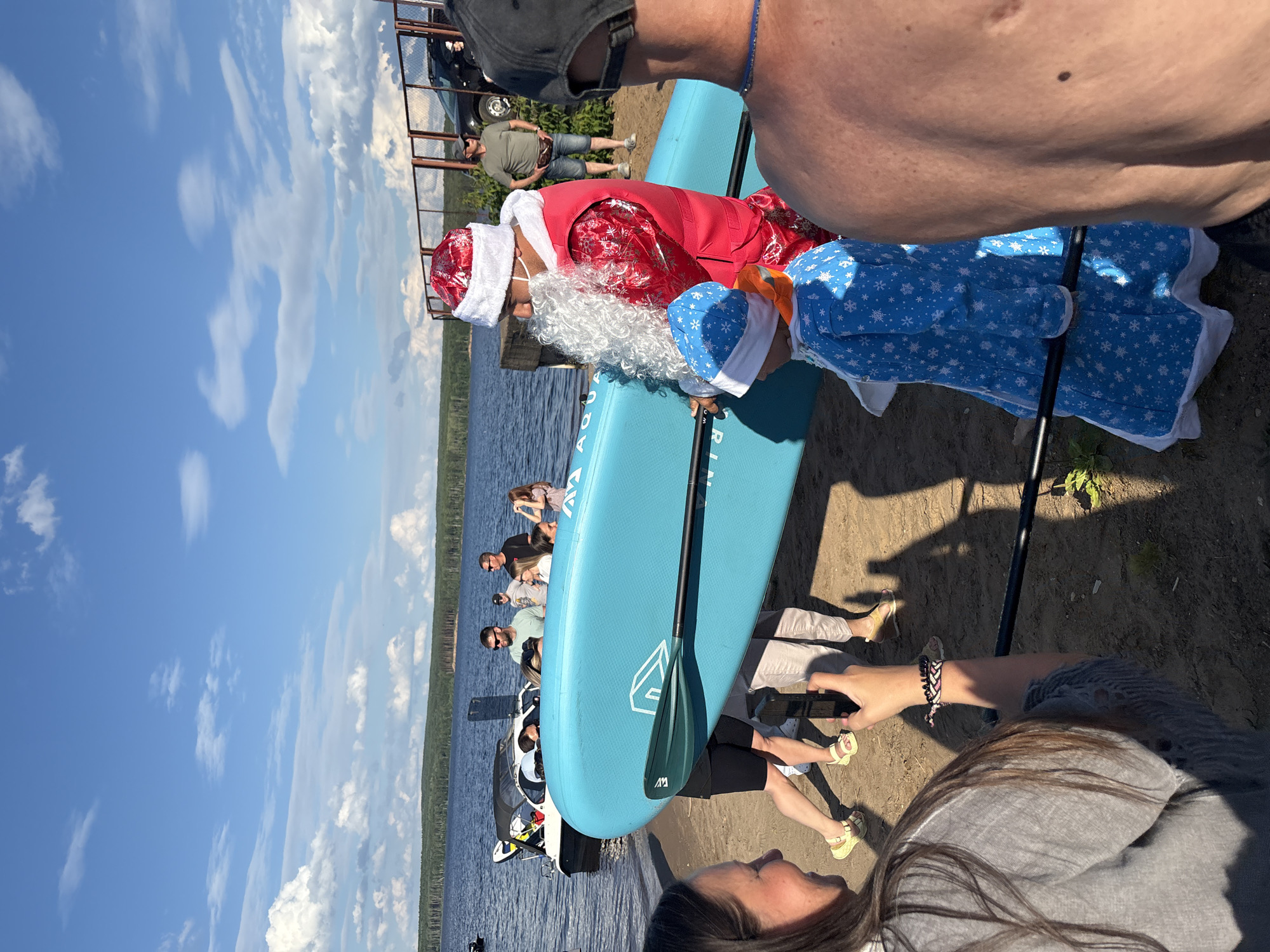 The image captures a group of people gathered on a sandy shore by a body of water, likely a lake or river, under a bright, cloudy sky. The scene appears to be set during the daytime, with strong sunlight casting shadows.
A prominent blue stand-up paddleboard lies on the sand. On the paddleboard, a person dressed in a Santa Claus costume, complete with a white beard and red hat, is seated. Another individual, possibly a child, is dressed in a blue garment with a snowflake pattern and a matching hat.
Several other people are visible in the background, some standing and observing. One person is holding a mobile phone, as if taking a picture or video. A boat is docked near the shore, and a metal railing structure is also present. The overall atmosphere seems relaxed and festive, with people enjoying an outdoor activity.
The visible text on the paddleboard is a logo that appears to be stylized letters within a diamond shape, and possibly the word "AQUA". The image is oriented vertically.