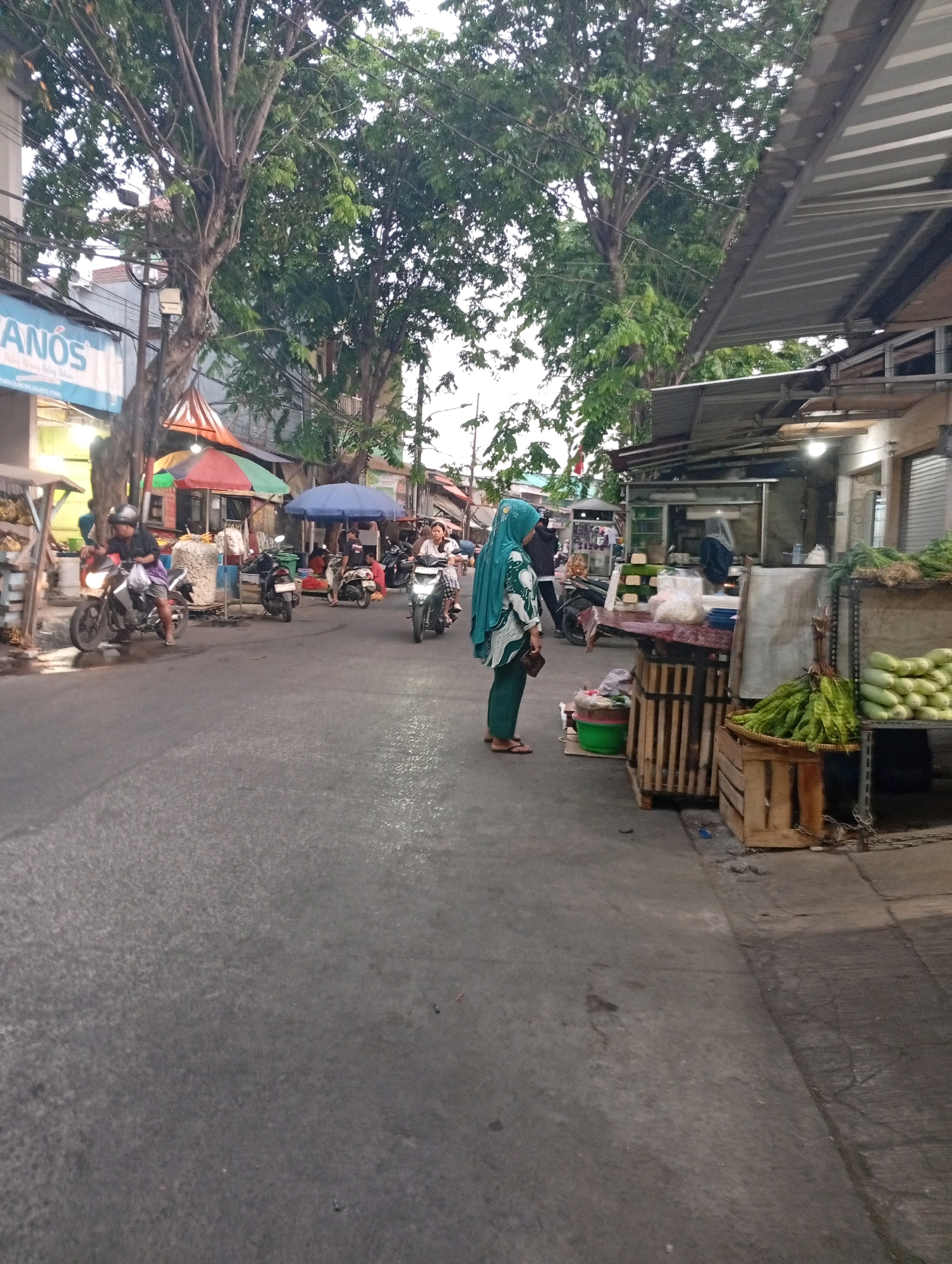 This is a street-level, medium-shot, vertical-format photograph taken in North Jakarta, Indonesia, likely a market area. A gray asphalt road occupies the foreground and midground. On the right side of the road, a market stall is visible, featuring shelves stocked with various produce, including green vegetables. A woman in a teal hijab and a patterned top stands facing the stall, looking down at the items. Behind her, a white awning extends over the stall. On the left side of the road, several motorcycles are parked and in motion. One man on a motorcycle, wearing a helmet and dark clothing, is moving away from the viewer. Further down the street, more market stalls with colorful umbrellas and awnings are set up. Trees with dense green foliage line the street, with utility poles and wires visible among the branches. The overall lighting suggests it is late afternoon or early evening.