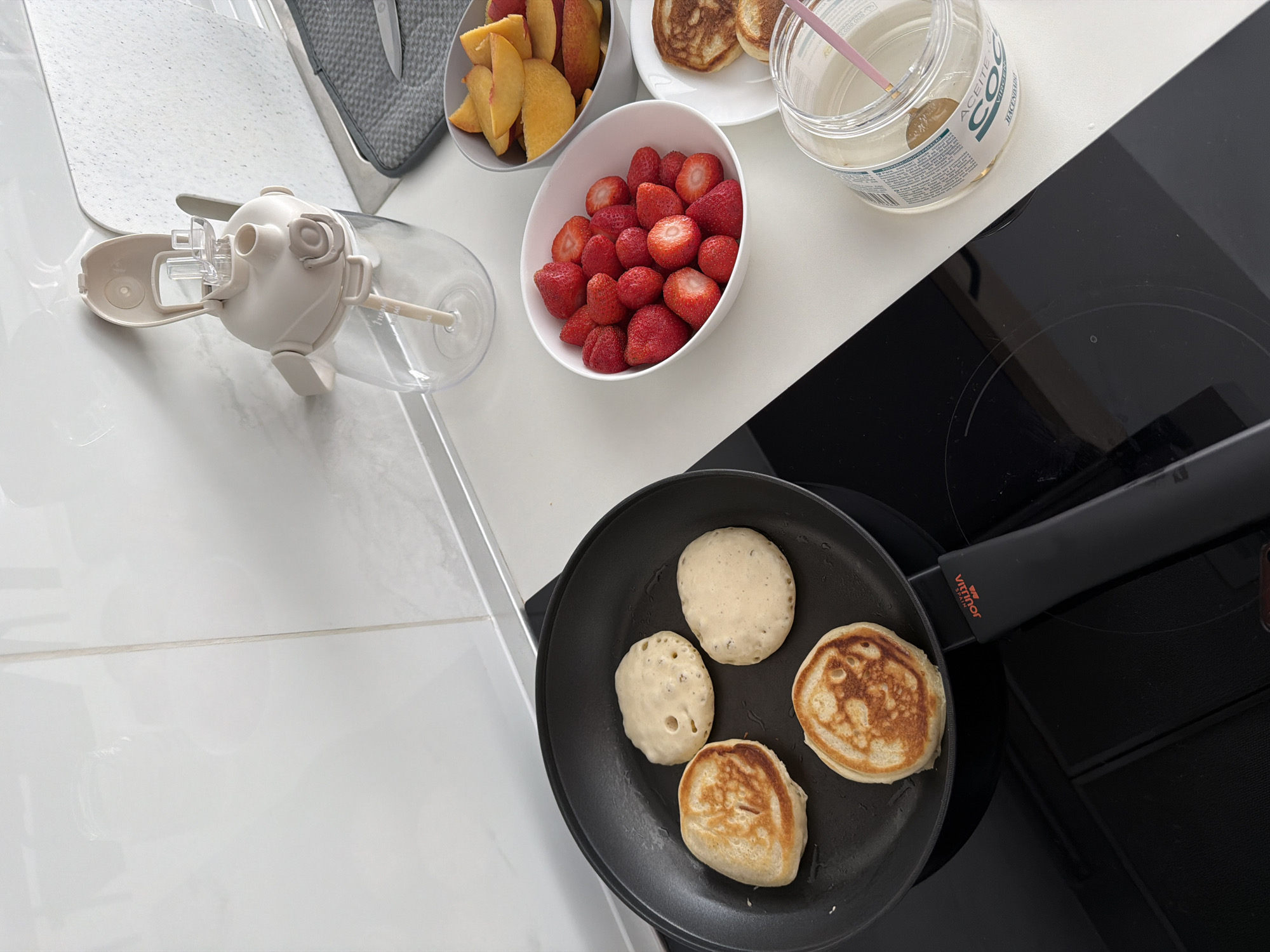 The image shows a kitchen scene in Finestrat, Spain, with a close-up view of pancakes being cooked on a stovetop. Four small, round pancakes are in a black frying pan, with two golden brown and two lighter in color, suggesting they are in different stages of cooking. To the right of the stovetop, there are three bowls filled with fruit: one with sliced peaches, another with fresh strawberries, and a third with a few cooked pancakes. A clear glass jar containing what appears to be coconut oil with a small brush inside is also visible. In the background, a white countertop and backsplash are present, along with a beige water dispenser with a straw. The overall impression is a breakfast preparation setting, possibly in a modern apartment or house.