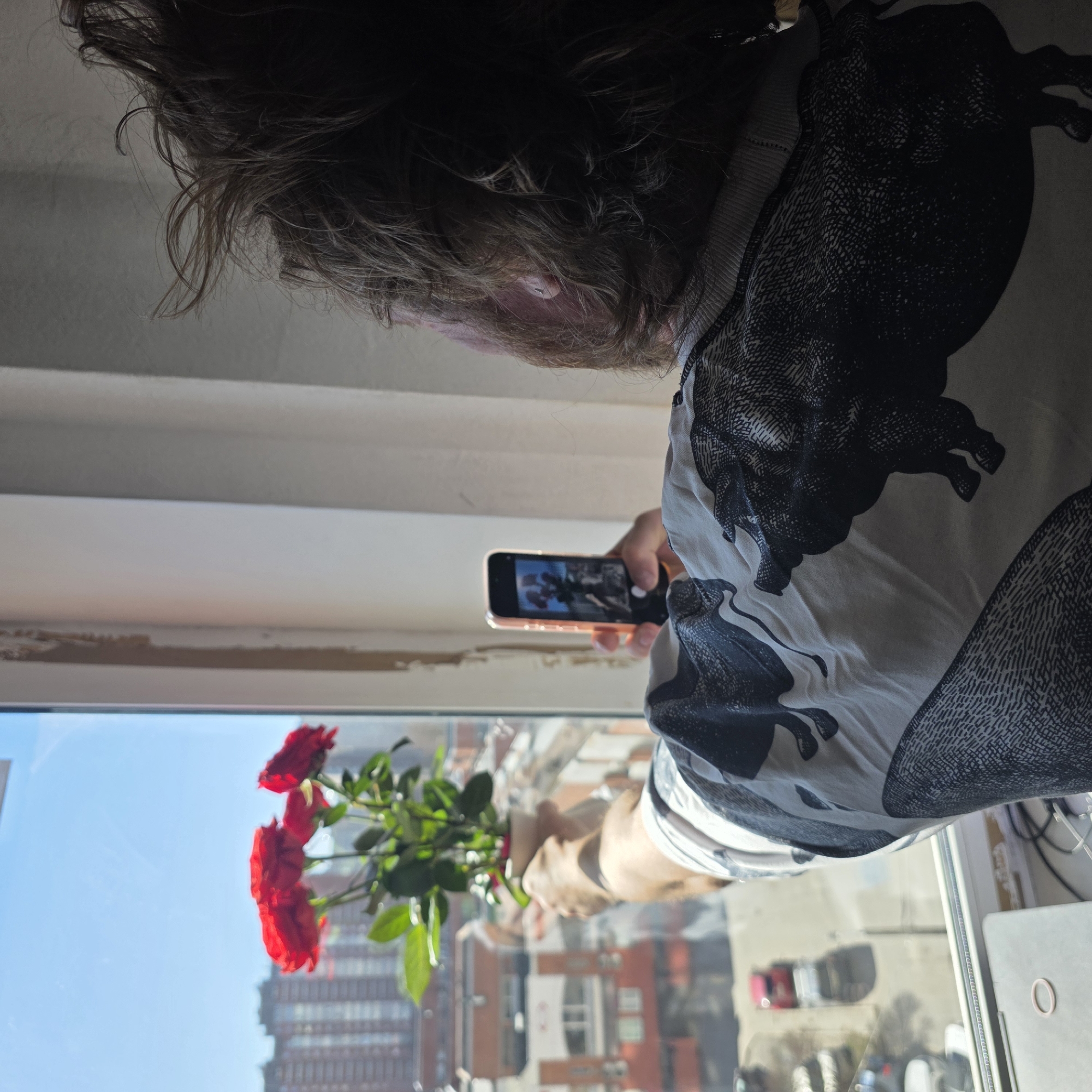 A person stands indoors by a window, facing the window and holding a bouquet of red roses. They are using a smartphone to take a photograph of the roses. The reflection in the window shows a cityscape, including a tall building resembling the Leader Tower in Saint Petersburg, Russia. The person has shoulder-length brown hair and is wearing a white shirt with a black pattern of what appears to be bulls. A window frame is visible, along with what appears to be an apartment or office window, including items on the window sill. The overall scene suggests someone is capturing a moment with flowers against the backdrop of the city.