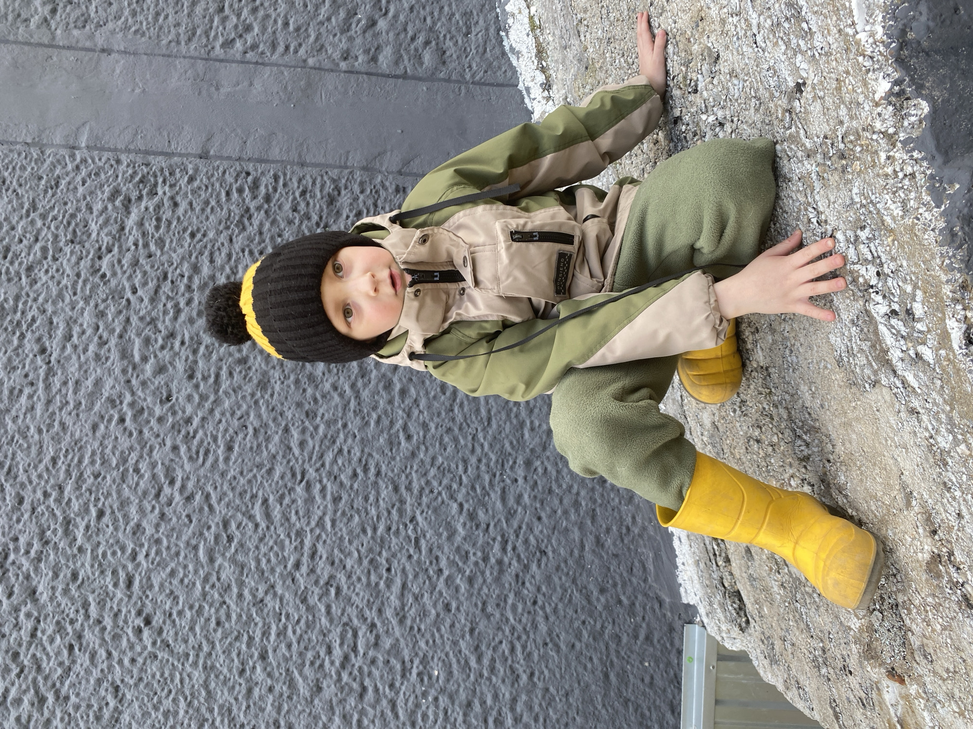 A young child, likely a toddler or preschooler, is captured outdoors in Noviy, Russia. The child is dressed in warm winter clothing, including a black and yellow striped beanie with a black pom-pom, a two-tone jacket featuring khaki/tan and olive green panels with multiple zippered pockets, olive green fleece pants, and bright yellow rain boots.
The child is seen sitting or crouching, looking directly at the camera with a calm and curious expression. Their hands are resting on a rough, light-colored surface to their right, which appears to be natural rock, ice, or snow-covered ground. To the child's left is a dark grey, heavily textured wall or vertical surface. A darker, possibly wet or icy, patch is visible where the two main background surfaces meet on the right. A silver metal object is partially visible at the bottom right of the frame.
The warm attire suggests cool or cold weather conditions. On a black tag above a zippered pocket on the jacket, the text "FORGEND" or "FORGJEND" is visible. The scene captures a moment of the child observing their surroundings in an outdoor environment.