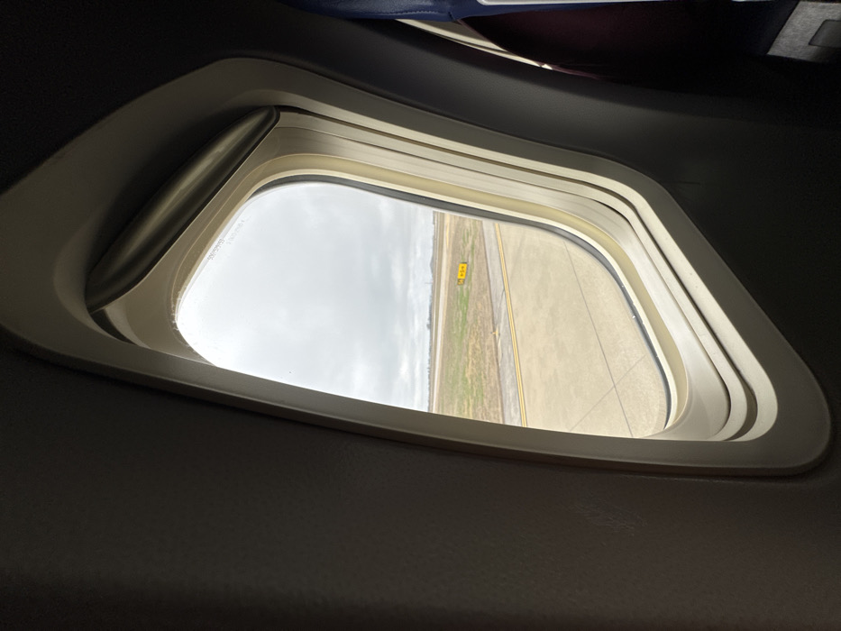 The image displays a view from inside an aircraft, looking out through a passenger window at an airport in Nashville, United States of America. The window features a light beige frame with a multi-layered appearance and a metallic element along its top edge, possibly part of a shade mechanism. To the immediate left, a dark grey interior panel of the aircraft is visible. To the right, a portion of a passenger seat back, appearing dark blue and maroon, is present. Outside the window, a concrete airport tarmac with parallel double yellow lines is visible in the foreground. Beyond the tarmac, a strip of dry, light green and brown grass extends. In the midground, a yellow square sign displaying "S6" in black text is positioned on the grassy area. The sky above is overcast and uniformly grey. The aircraft appears to be stationary on the ground. No individuals are visible within the frame.