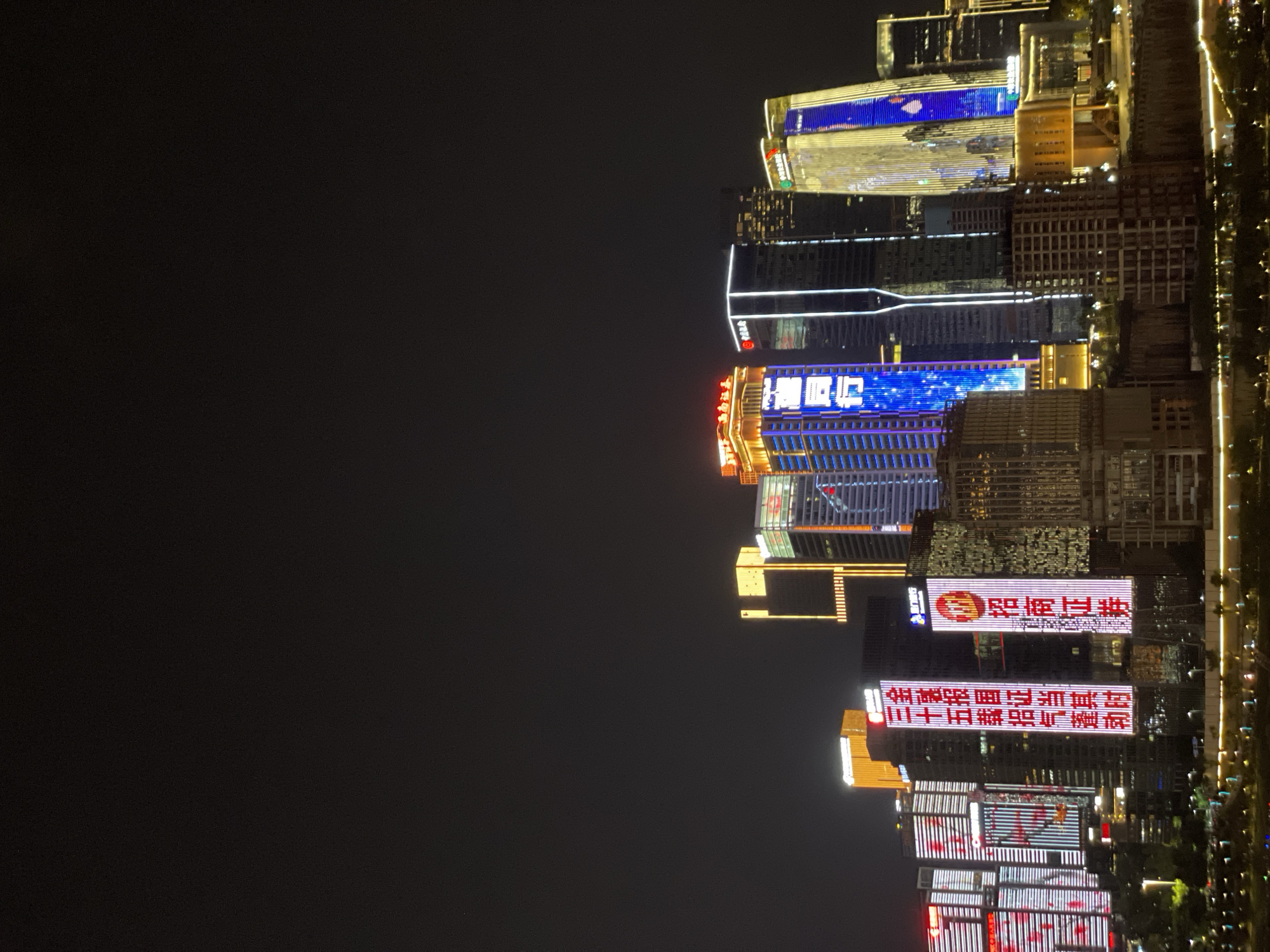 This media file shows a nighttime scene of a city skyline, likely in the Yuzhong District of China. The image is vertically oriented, with a dark sky dominating the left half and a brightly lit cityscape on the right. Several modern skyscrapers are visible, adorned with a multitude of lights and illuminated signage.
The buildings display a variety of architectural styles, with some featuring sleek glass facades and others with more textured, grid-like patterns. The lights on the buildings are diverse, ranging from white and blue to orange and red, creating a vibrant and dynamic urban landscape.
Prominent text is visible on several of the buildings. One sign, illuminated in red and white, reads "招商证券" (Zhaoshang Securities). Below this, another large sign in red characters says "金工报国证当其时 三十五载招气蓬勃" (The golden industry serves the nation when the time comes, thirty-five years of vigorous spirit). Other buildings feature vertical and horizontal strips of light, some in red and white patterns that resemble Chinese lanterns, and others with blue light displays.
The scene appears to be captured from a distance, possibly across a body of water, given the faint reflections of lights on what could be a river or bay along the bottom edge of the cityscape. The time of day is clearly night, with the artificial lights of the city standing out against the darkness. The weather seems clear, allowing for sharp visibility of the illuminated structures. No people are discernible in the image, focusing the attention solely on the architectural display of the city at night.