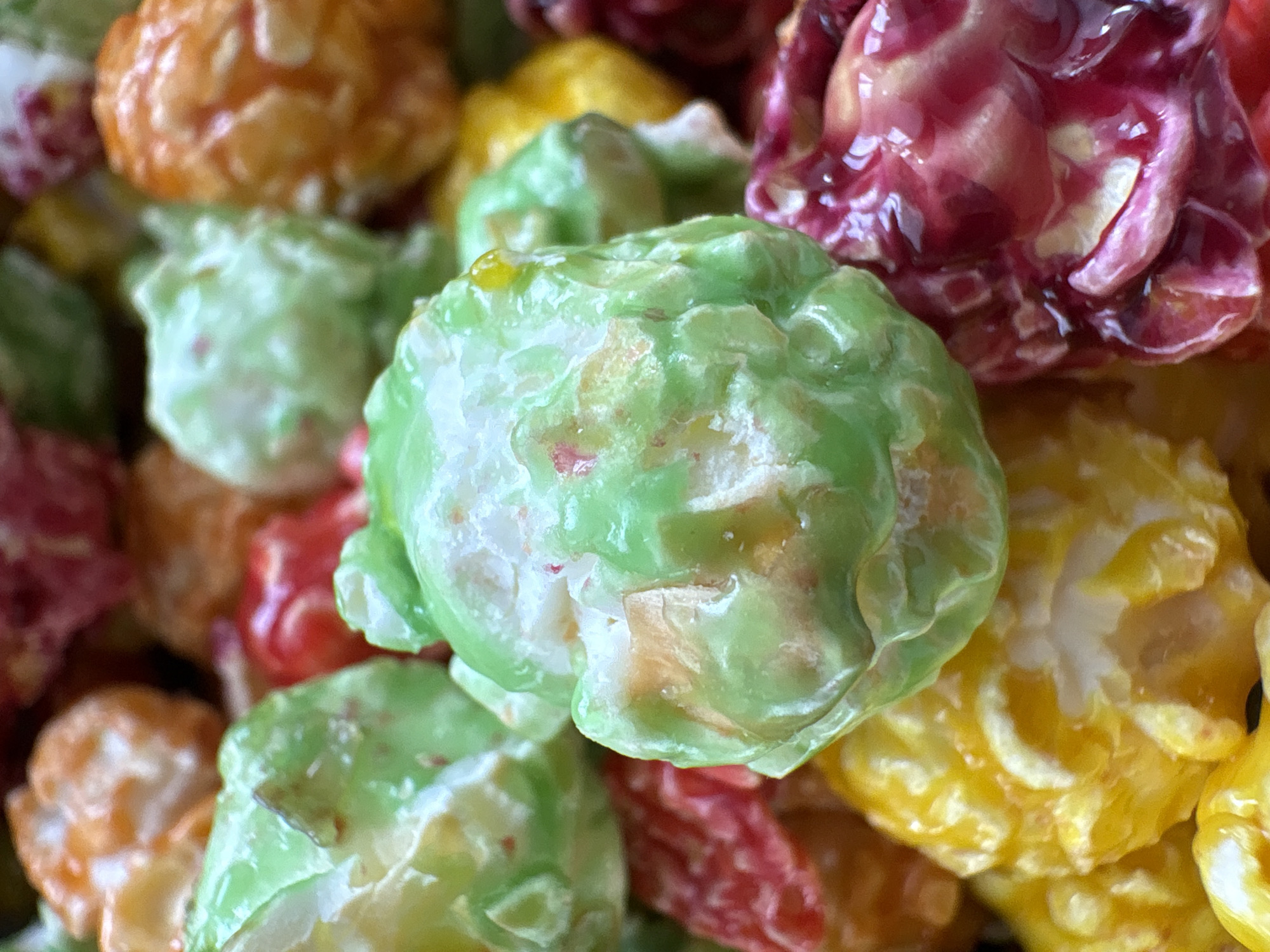 A close-up shot shows several pieces of colorful candy-coated popcorn. The popcorn pieces are green, orange, yellow, and red. The background is out of focus. There is no evidence the photo was taken in Chaikovsky, Russia. There are no people visible in the photo.