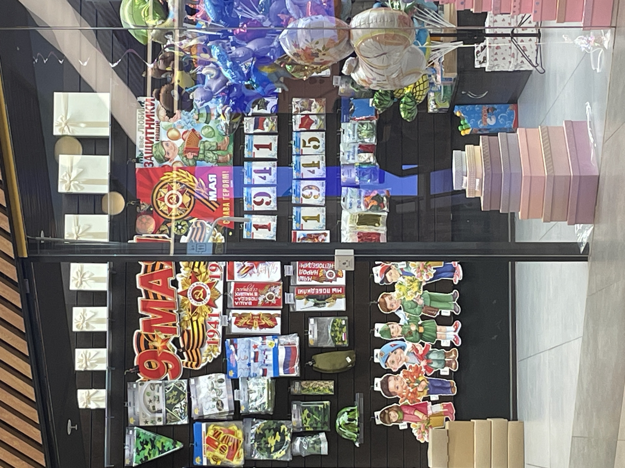 The image depicts a display case filled with various items, likely for sale, in what appears to be a shop or store, potentially located in Moscow, Russia. The items relate to Victory Day, a Russian holiday commemorating the victory over Nazi Germany in World War II. The display includes decorations like balloons, banners, and stickers. Some of these items feature the years 1941 and 1945, key dates in the war. There are also images of soldiers, military equipment, and patriotic slogans, like "We love you, defenders of our land," and "Our cause is just, we will win!" Gift boxes are also displayed. The scene suggests a commercial setting preparing for or celebrating this significant holiday. The photograph's perspective is slightly rotated.