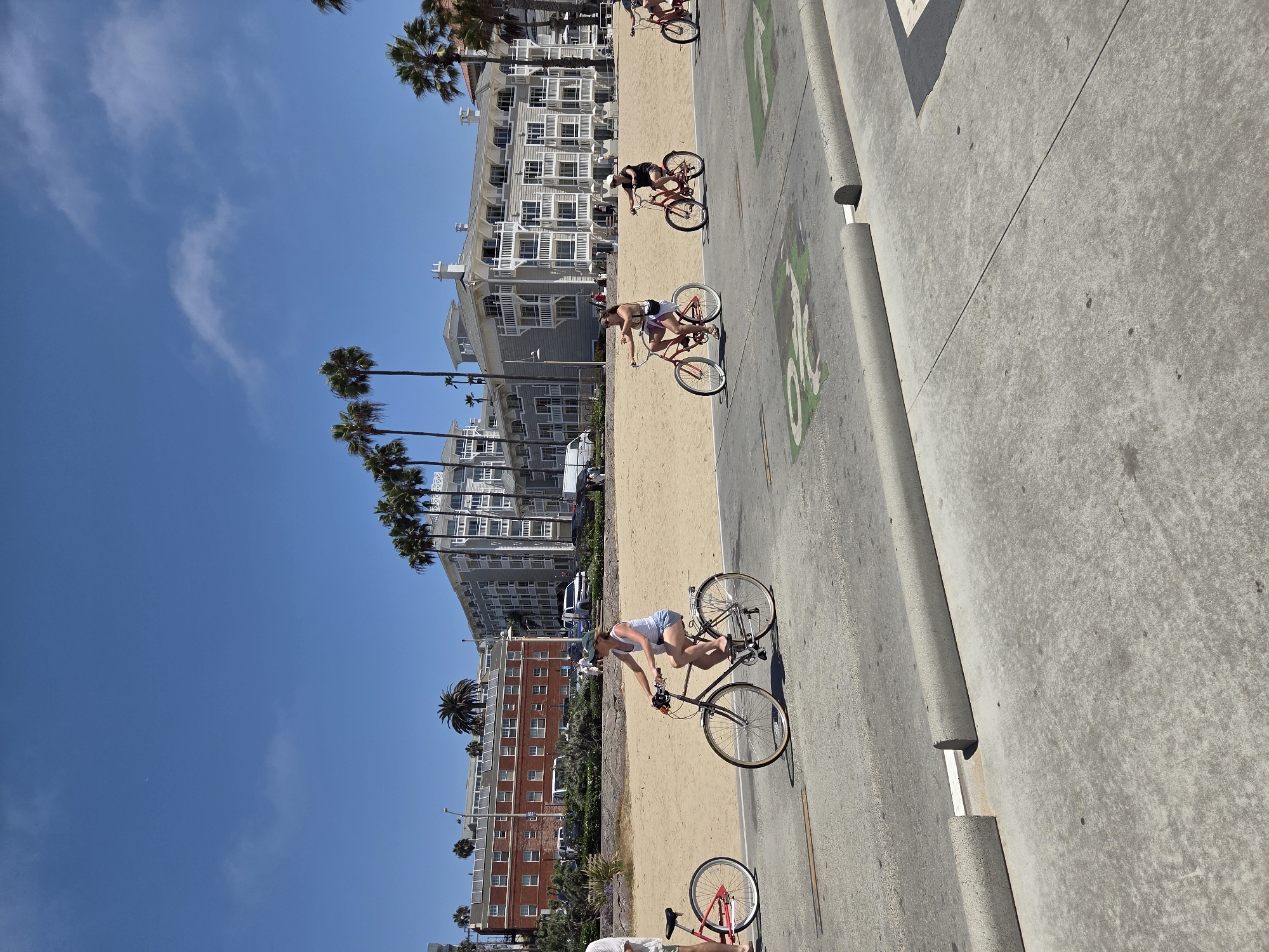 In Santa Monica, California, along a paved beachfront path adjacent to a sandy beach, several individuals are depicted riding bicycles. The scene is set on a bright, sunny day with a clear blue sky. In the background, a row of multi-story buildings, likely apartments or hotels, with light-colored facades and balconies, are visible, interspersed with palm trees. A low wall separates the paved path from the sand. One person, wearing a white tank top and denim shorts, is riding a dark-colored bicycle in the foreground, facing right. To their left, another person on a red bicycle is partially visible. Further down the path, three more individuals are riding bicycles, all facing right. The paved path has green painted symbols, possibly indicating a bike lane or directional markers. The overall impression is a leisure activity taking place in a coastal urban environment.