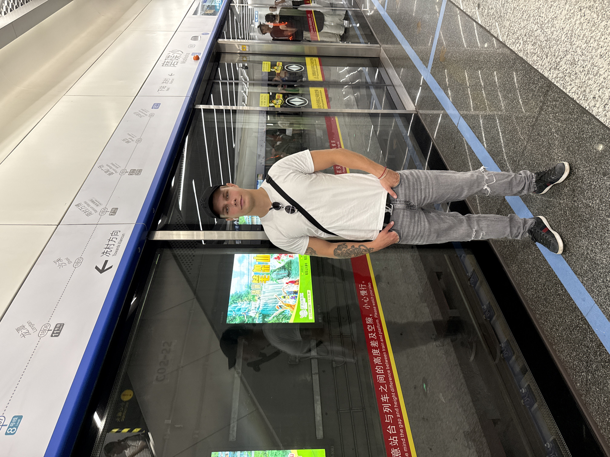 A man stands inside the Modiesha station in Haizhu District, China. He is wearing a white t-shirt, grey ripped jeans, a black baseball cap, and black and red sneakers. He has a black bag slung over his shoulder and sunglasses attached to the bag strap. The background shows the station platform and train doors with safety signage in Chinese and English, and route maps for the Guangzhou Metro system.