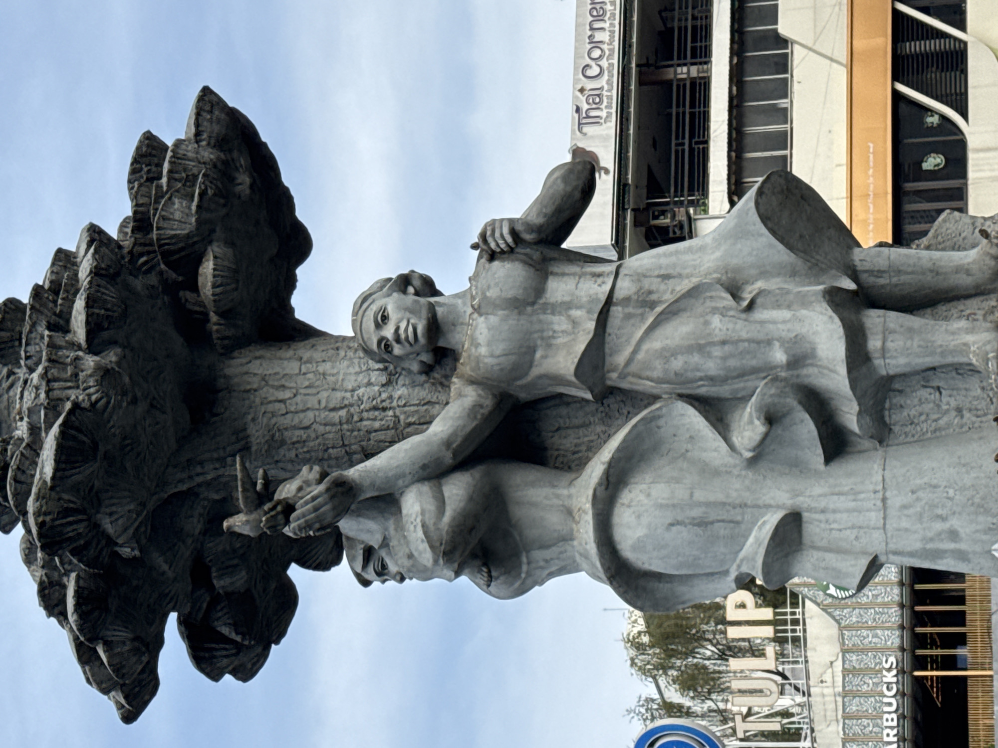 The image shows a large stone sculpture of two women standing next to a tree trunk. The woman on the right is holding a bird in her outstretched hand. Behind the sculpture, buildings are visible, including a building with a sign that reads "TULIP" and another with a sign that reads "Thai Corner". A Starbucks logo is also visible on one of the buildings. The sculpture is located in the city of Da Lat, Vietnam, a popular tourist destination known for its French colonial architecture and lush landscapes. The sculpture appears to be a monument or public art installation, possibly representing peace or nature. The sky is cloudy, suggesting an overcast day.