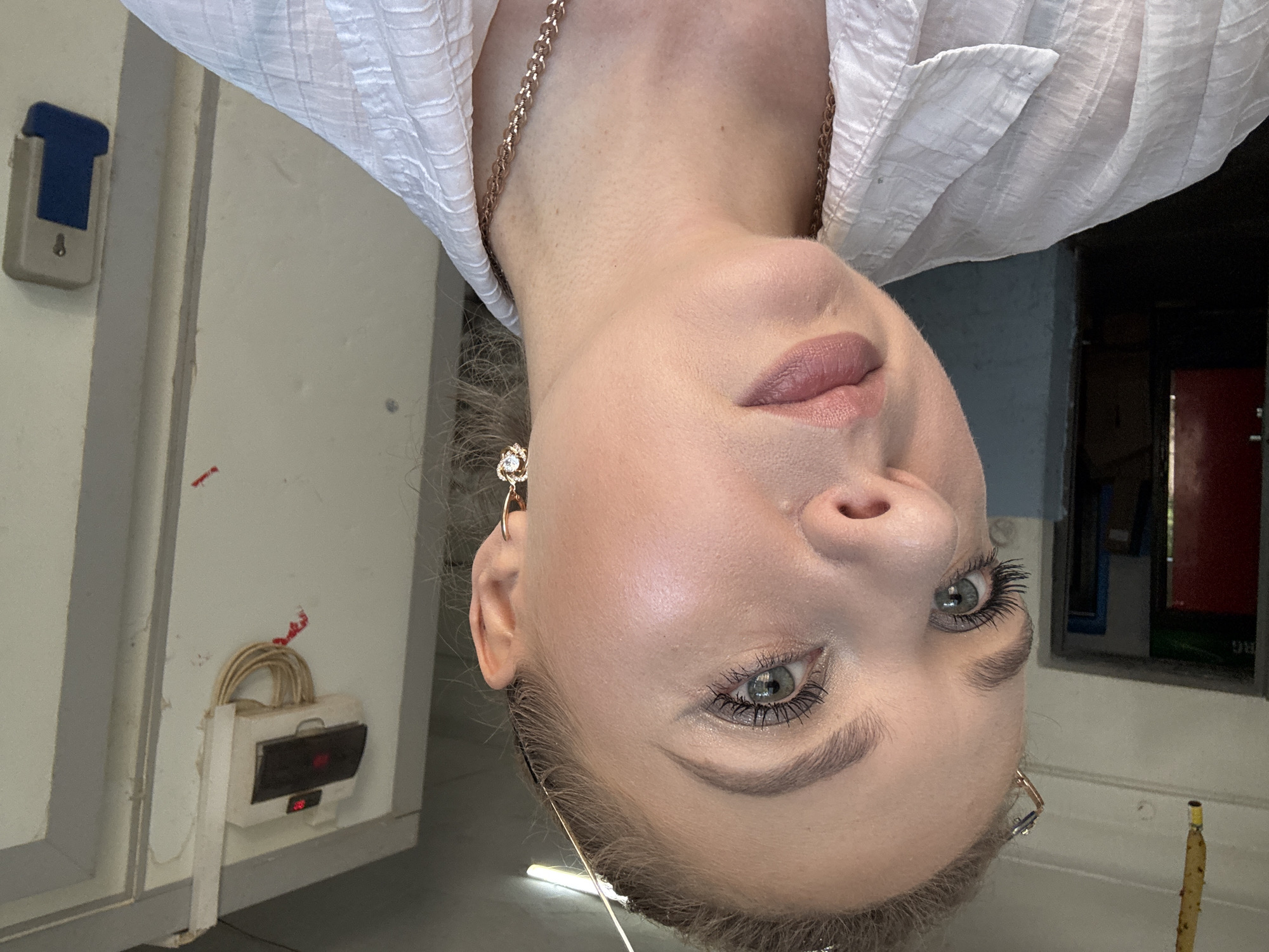 A close-up, outdoor, medium shot shows a young, Caucasian woman from the chest up, facing forward and slightly to her left. The woman has blonde hair pulled back, and is wearing a white, textured shirt, gold earrings in the shape of hearts with clear stones, and a gold necklace. Her makeup includes mascara, eyeliner, eyeshadow, and light pink lipstick. Her left eye is open and looking toward the camera, while her right eye is partially visible.
The background is out of focus and appears to be an urban environment. To the left, a dark doorway is visible with a red door. In the center background, a white electrical panel is mounted on a wall, with wires hanging below it. To the right of the electrical panel, a light-colored door or wall is visible. The lighting suggests it is daytime.