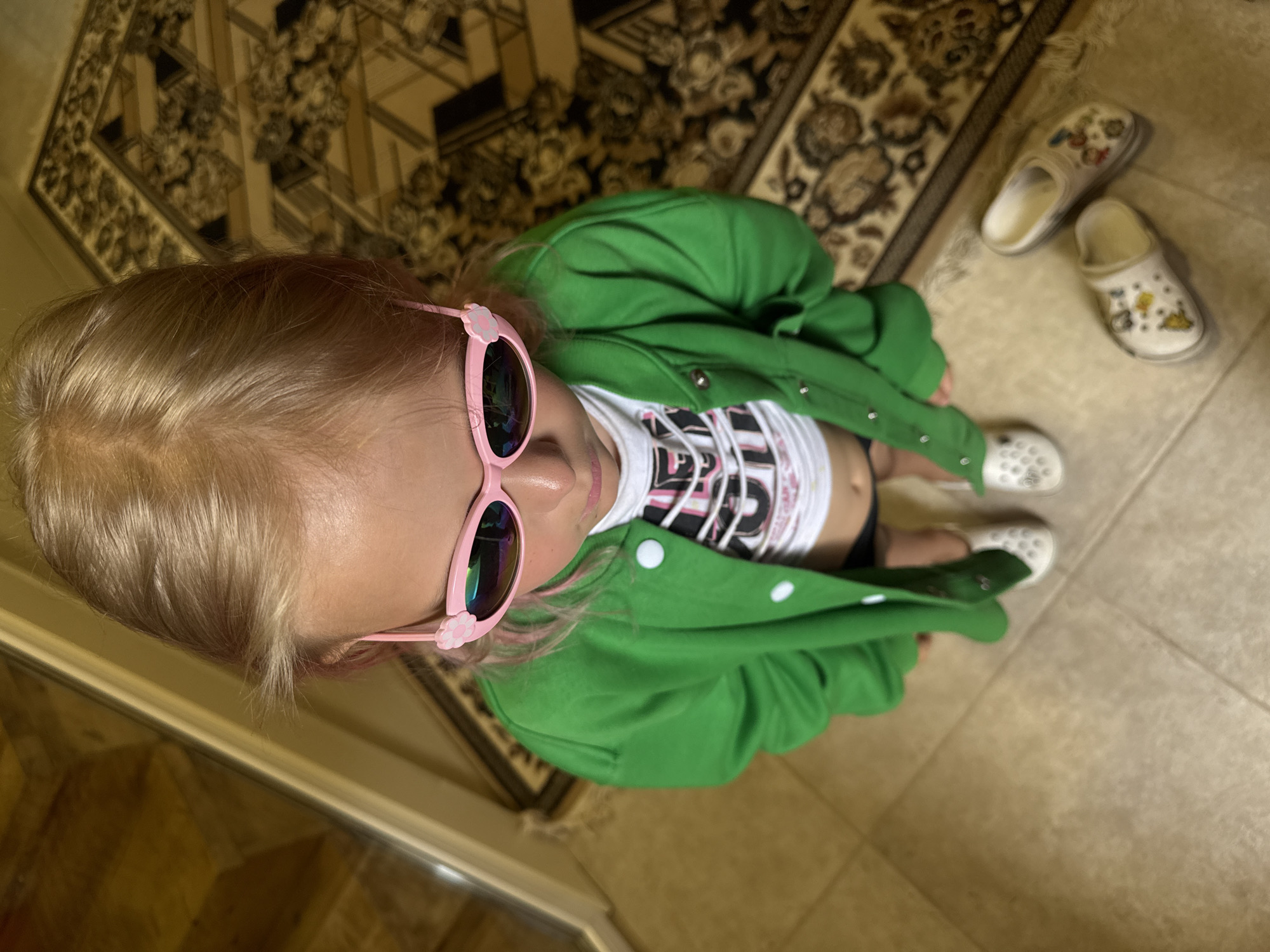 An overhead shot shows a young Caucasian girl standing on a tiled floor next to a patterned rug. The girl, with blonde hair and pink highlights, is wearing pink sunglasses with flower details, a green jacket, and a white t-shirt. Her feet are clad in white Crocs. Two additional pairs of decorated white Crocs lie on the rug near her feet. The background includes a wooden door frame.