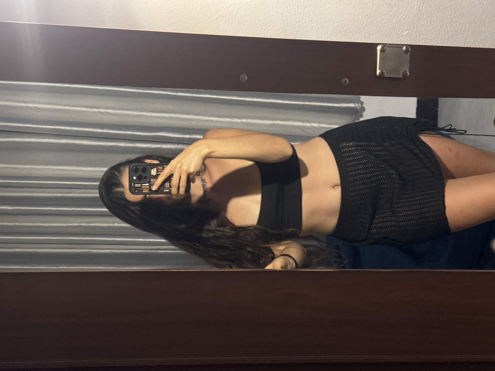 A young woman is lying on a bed in a room, taking a mirror selfie. She is wearing a black crop top and a black, possibly crochet, mini skirt. The background consists of a grey curtain and white walls. The reflection in the mirror shows part of the room's interior, including the bed and the headboard of a bed. The image was most likely taken indoors in a residence; there's no explicit information linking it to Mũi Né, Vietnam.