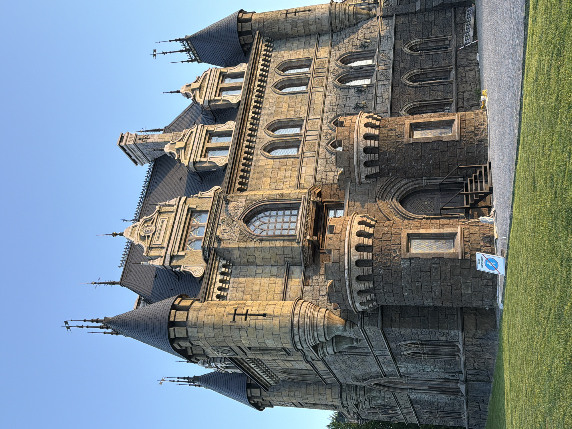 The image shows Garibaldi Castle in Khryashchevka, Russia. It is a neo-Gothic architectural structure constructed of stone with towers, spires, and arched windows. A grassy lawn is present on the right side of the image, and a paved path is visible adjacent to the grass. The sky is clear and blue. A sign is visible on the side of the castle and a set of outdoor stairs lead up to one of the doors.