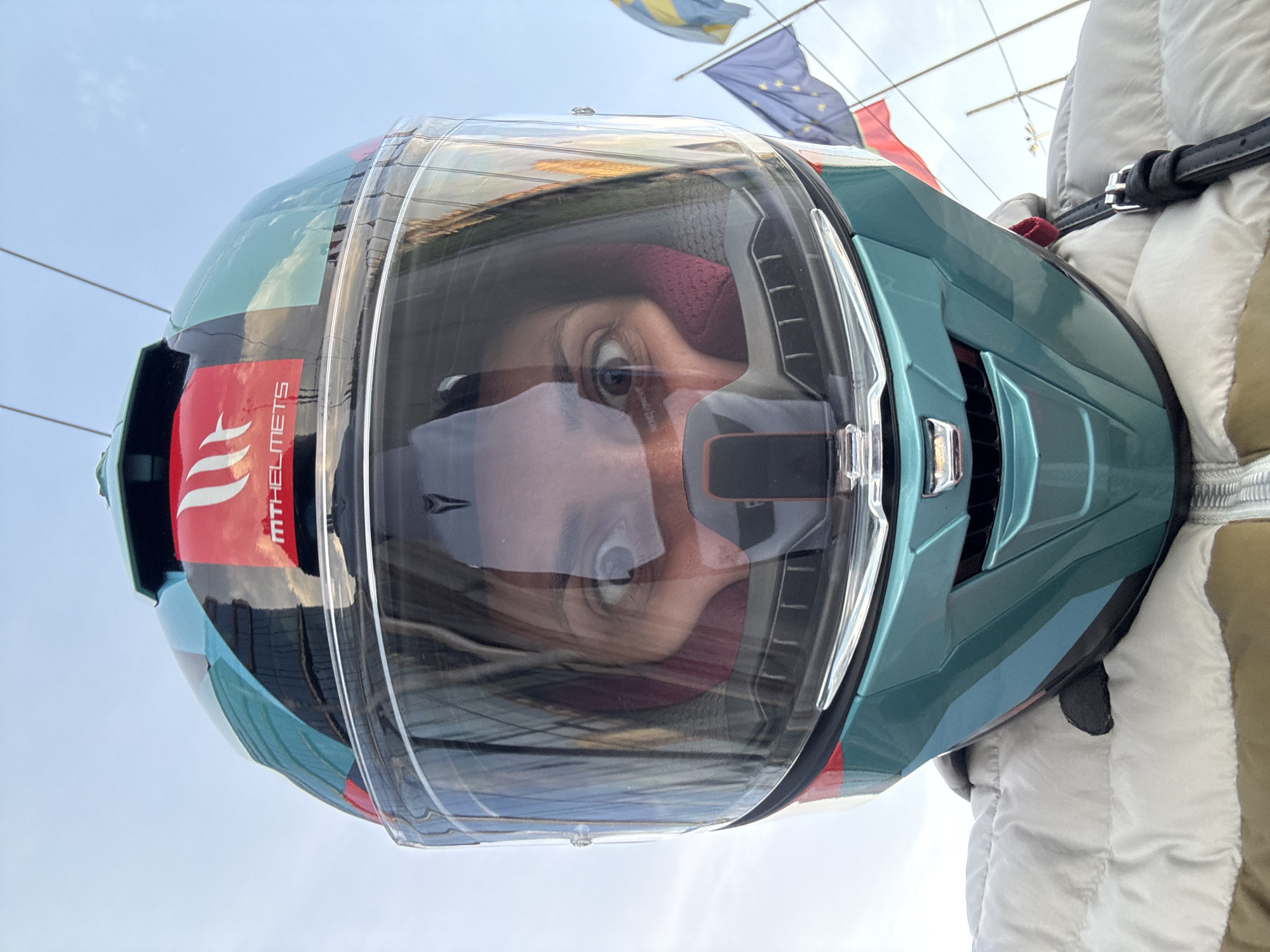 A low-angle shot depicts a person wearing a teal and blue MT Helmets motorcycle helmet. The clear visor reflects the person's face. In the background, partially visible, are flags, likely including the European Union flag, suggesting a governmental or official location. A portion of a white winter jacket is seen at the lower right. The photo was taken outdoors, with the sky visible in the background. The setting is likely the city of Sofia, Bulgaria, judging by the flag of Sofia, Bulgaria (yellow with a white lion) in the background.